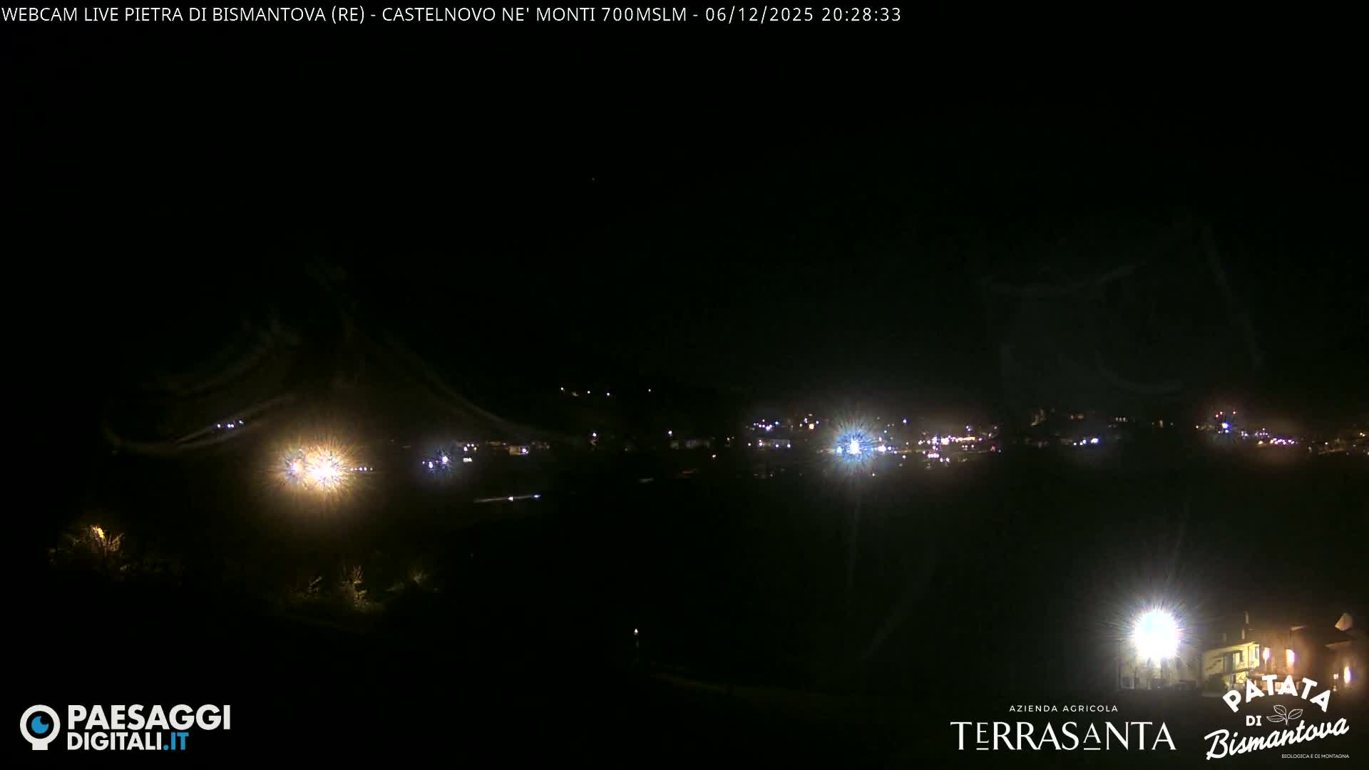 A dark night panorama shows a sprawling settlement illuminated by numerous bright lights, including prominent streetlights that create a distinct glow and lens flare effect, all under a clear sky.