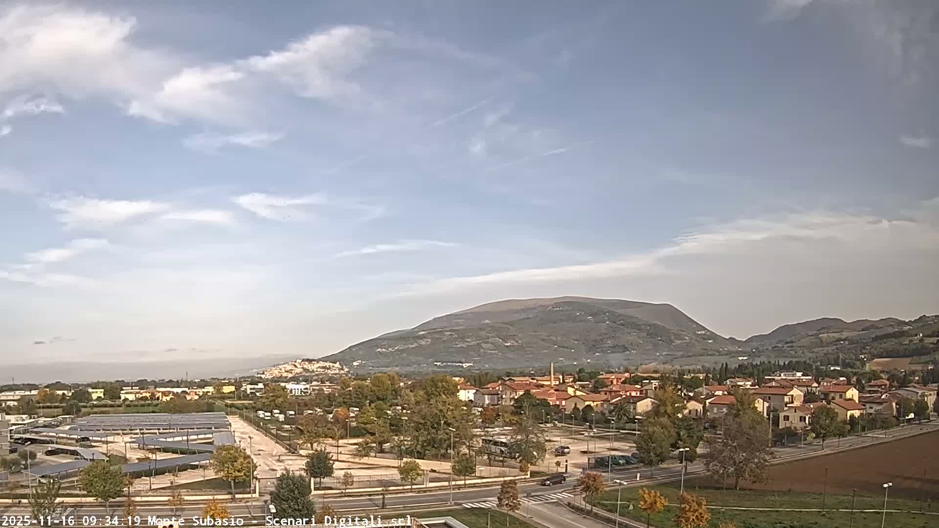 Under a clear and sunny sky with scattered white clouds, an expansive outdoor scene reveals a town with residential buildings, roads with vehicles, a prominent solar panel installation, and trees showing autumn colors, all framed by distant mountains.