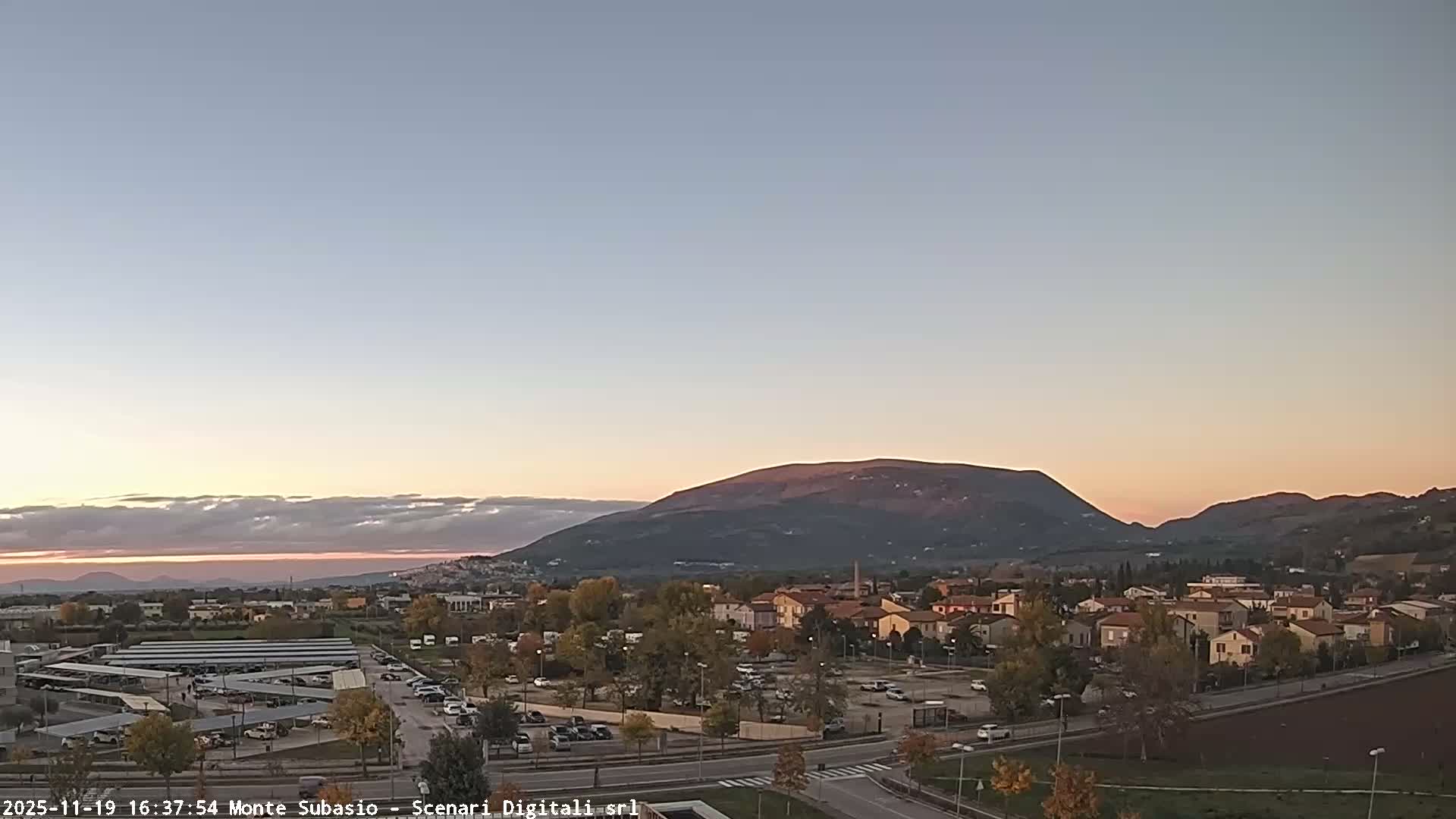 Under a clear and sunny sky with scattered white clouds, an expansive outdoor scene reveals a town with residential buildings, roads with vehicles, a prominent solar panel installation, and trees showing autumn colors, all framed by distant mountains.