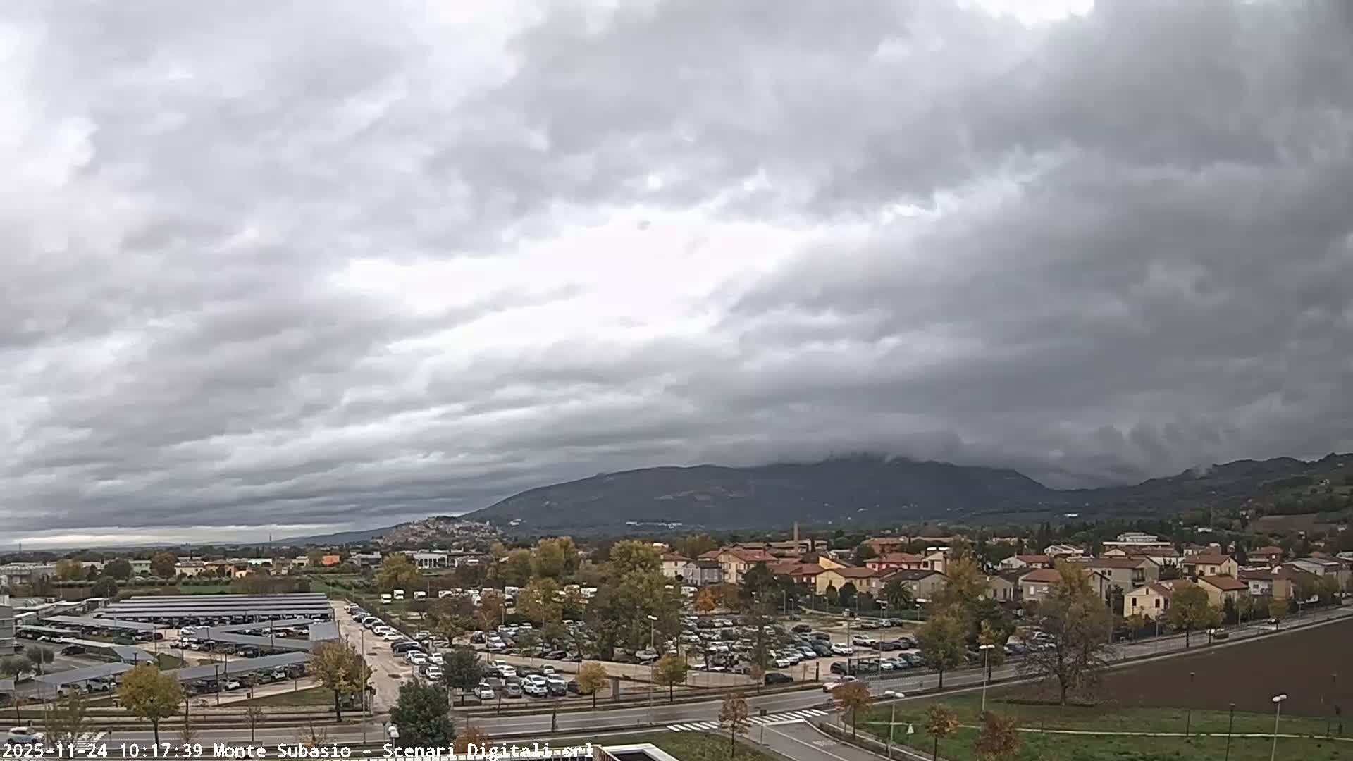 Under a heavily overcast and gloomy sky, a town with extensive parking lots, residential buildings, and autumn trees stretches towards distant, cloud-shrouded mountains.