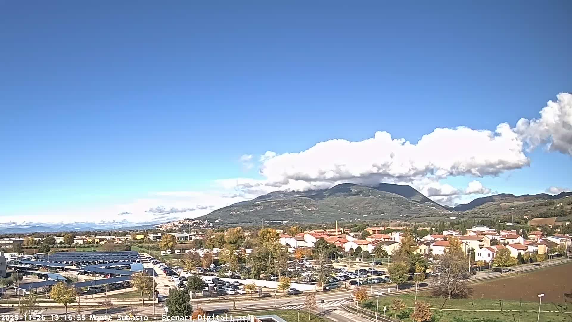 Under a heavily overcast and gloomy sky, a town with extensive parking lots, residential buildings, and autumn trees stretches towards distant, cloud-shrouded mountains.