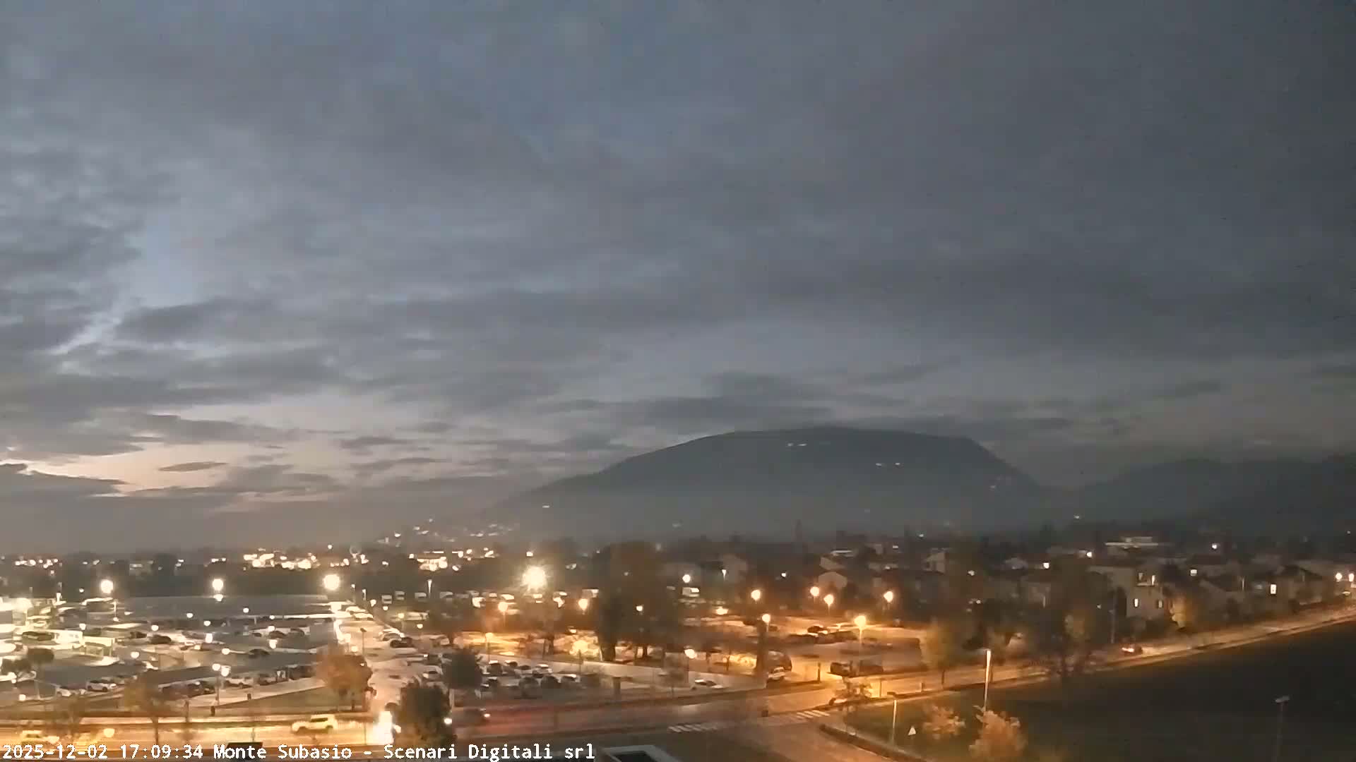 An overcast dusk settles over a town, its streets and parking lots illuminated, leading to hazy mountains under a dark, cloudy sky.