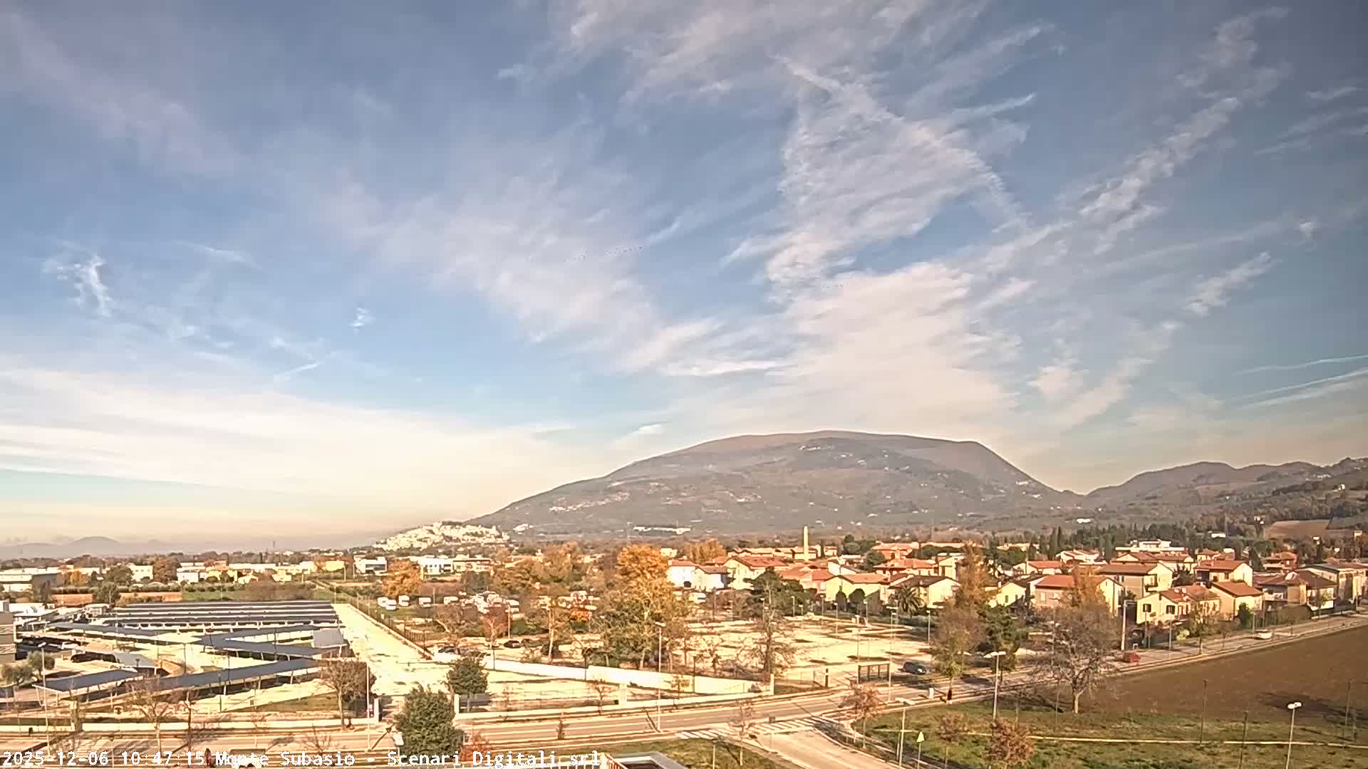 An expansive outdoor view reveals a town featuring red-roofed buildings, roads, and a large solar panel array amidst autumnal trees, set against a backdrop of rolling hills and a prominent mountain under a bright, partly cloudy sky.