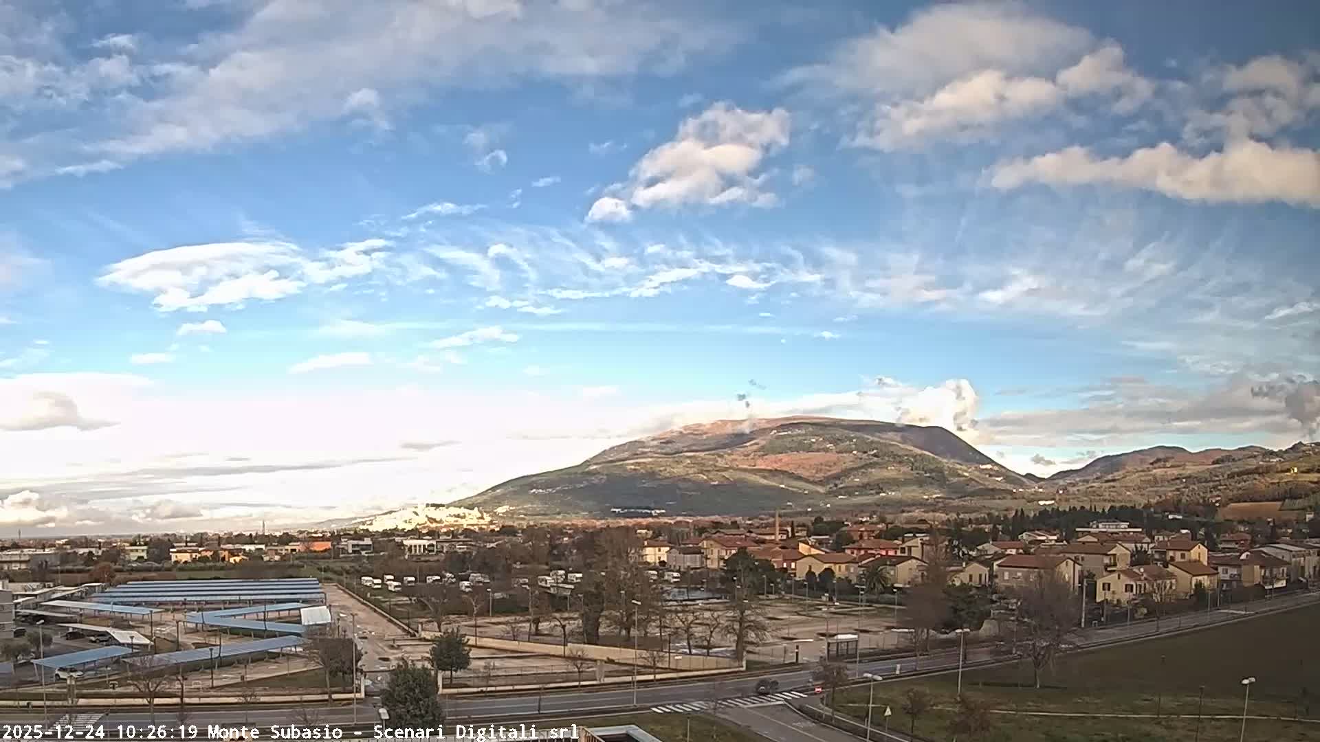An expansive outdoor view reveals a town featuring red-roofed buildings, roads, and a large solar panel array amidst autumnal trees, set against a backdrop of rolling hills and a prominent mountain under a bright, partly cloudy sky.