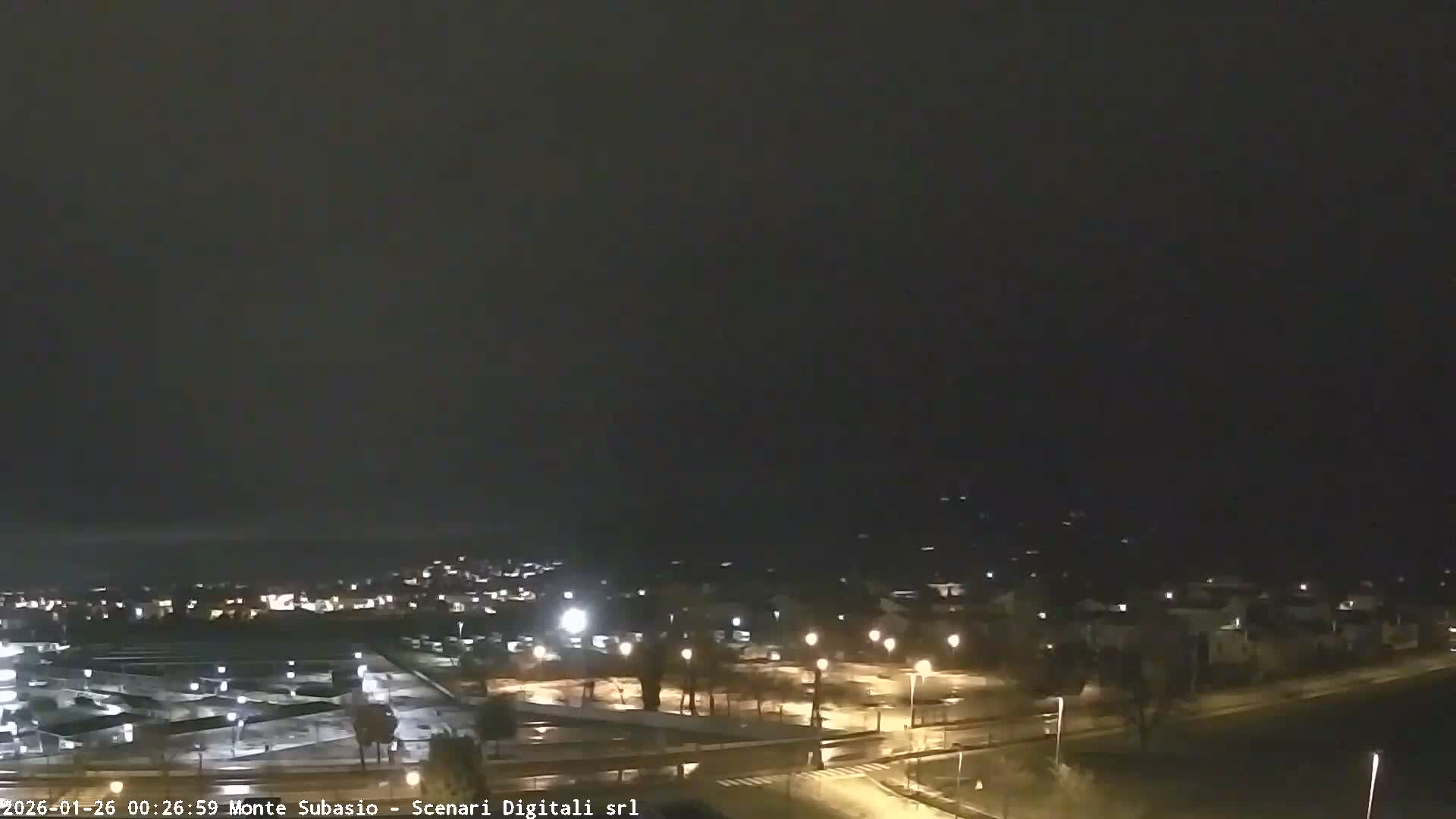 A nighttime view of a town illuminated by streetlights, with wet ground reflecting lights under a dark, cloudy, and rainy sky.