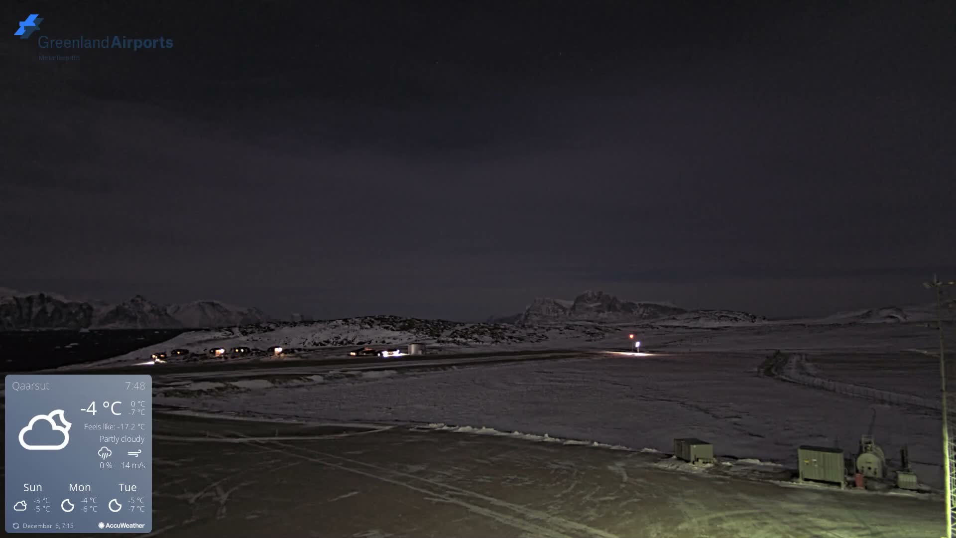 The image displays a snow-covered airport tarmac and runway with distant illuminated buildings and dark, snowy mountains under a dark, partly cloudy sky, indicating cold winter conditions.