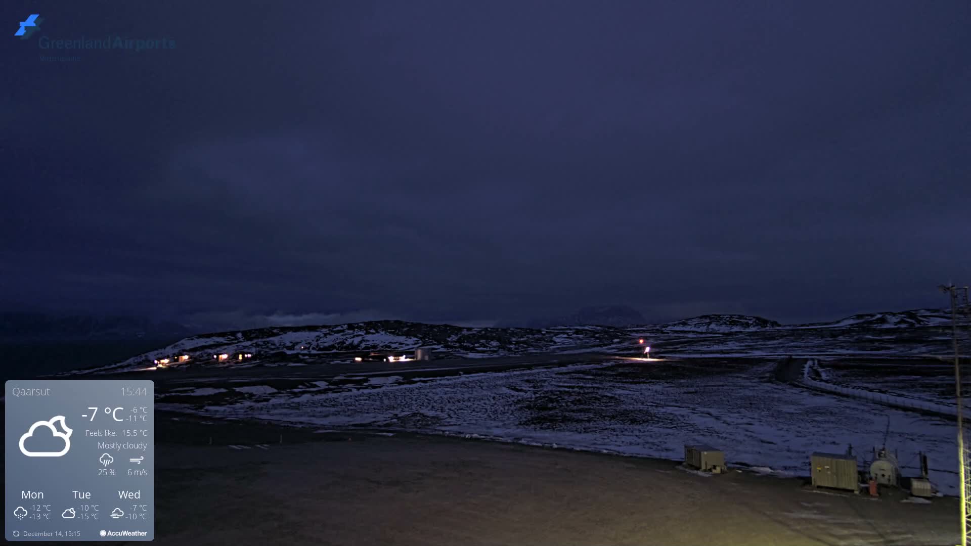 The image displays a snow-covered airport tarmac and runway with distant illuminated buildings and dark, snowy mountains under a dark, partly cloudy sky, indicating cold winter conditions.