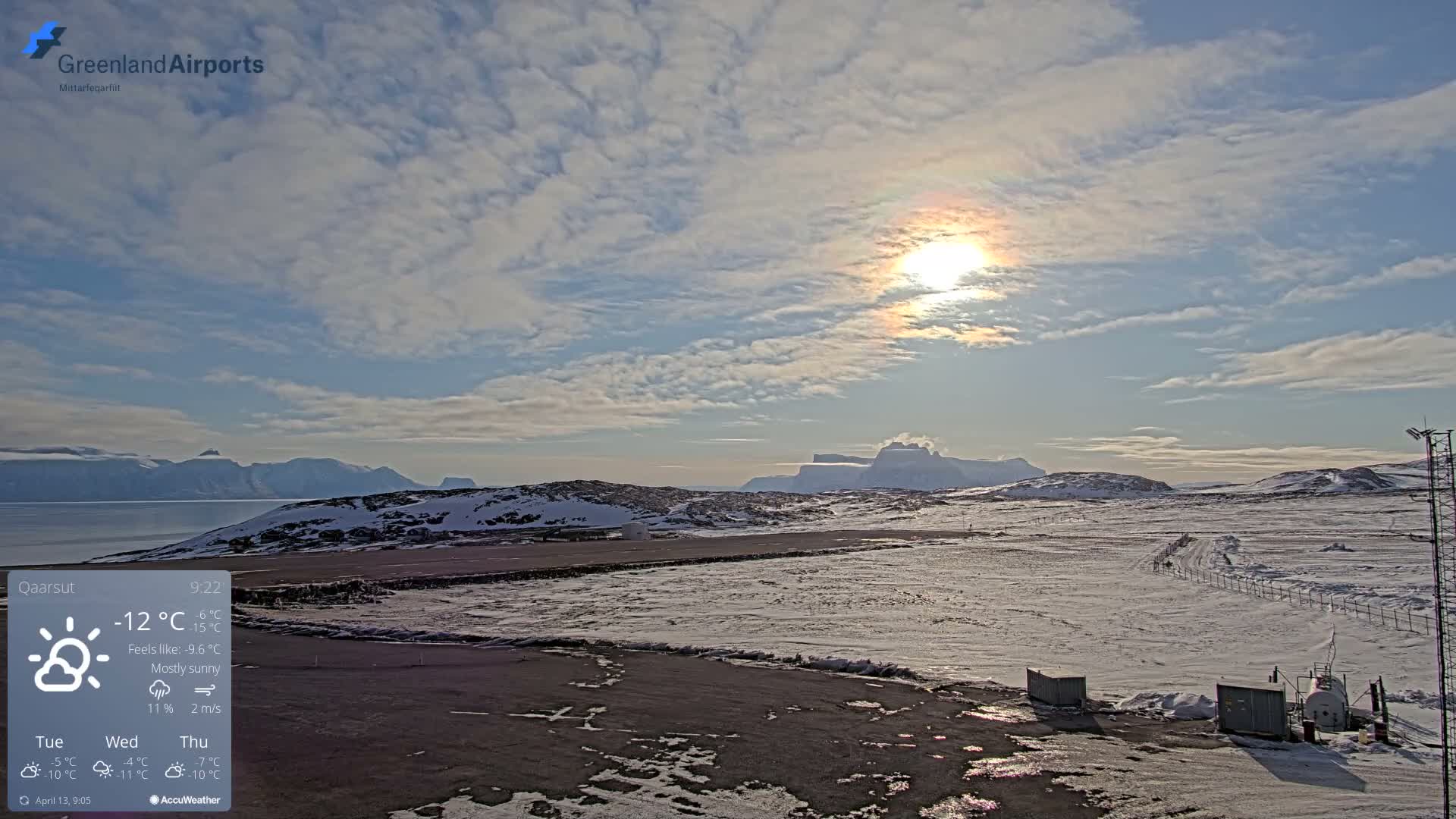 The image displays a snow-covered airport tarmac and runway with distant illuminated buildings and dark, snowy mountains under a dark, partly cloudy sky, indicating cold winter conditions.