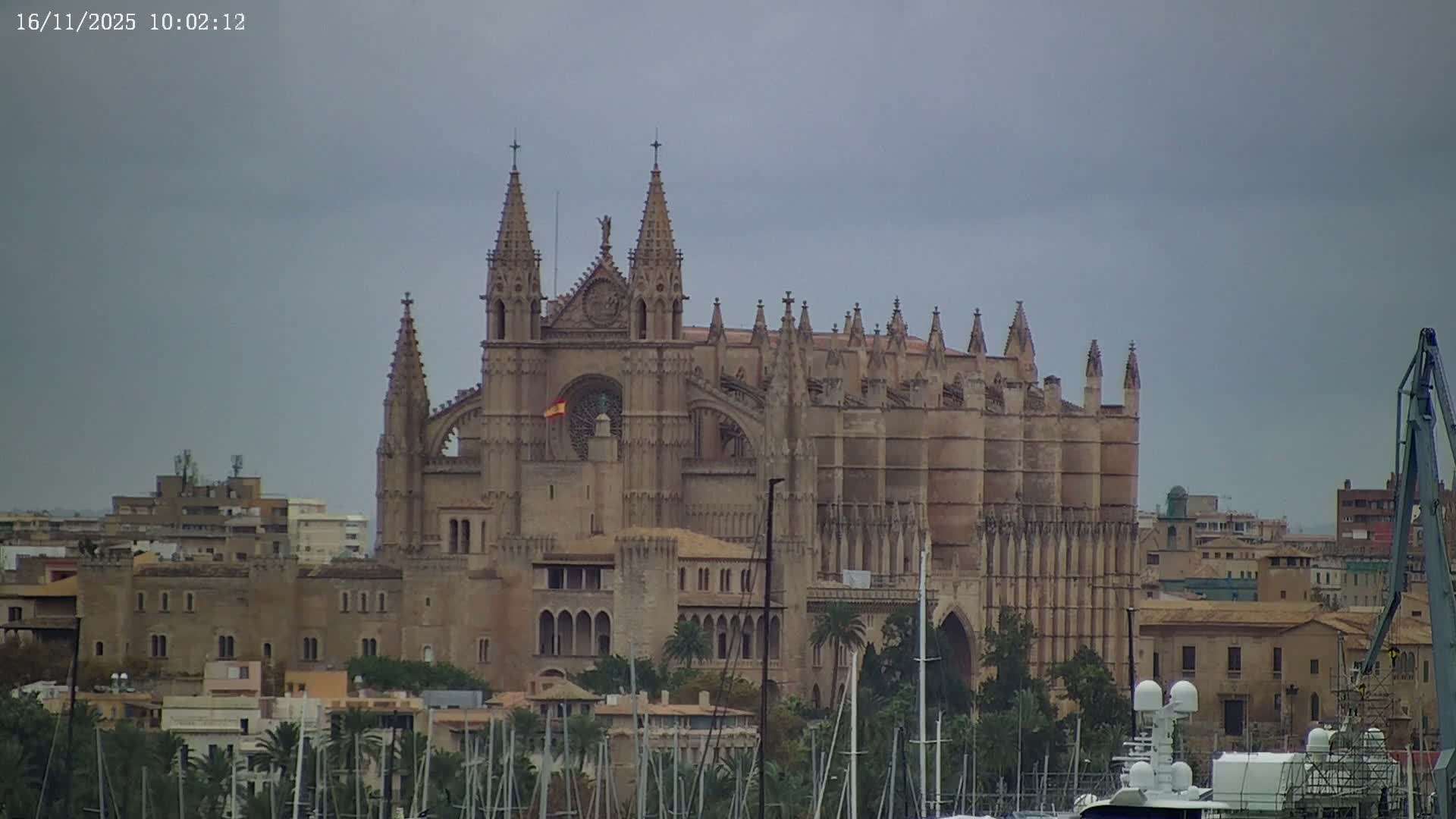 A majestic Gothic cathedral overlooks a bustling marina with numerous sailboats and yachts, surrounded by city buildings and palm trees, all beneath a grey, overcast sky.
