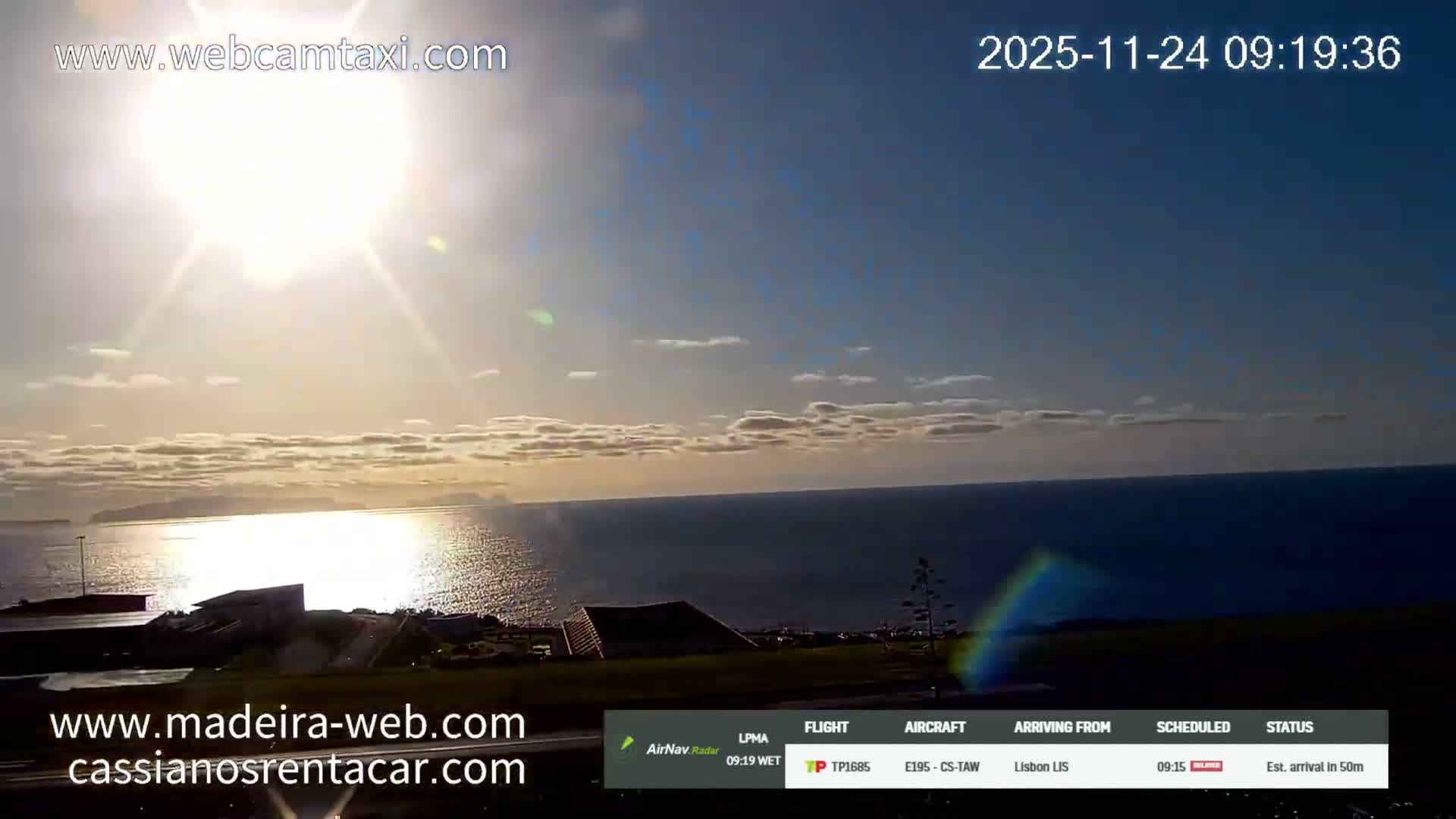 A bright and sunny morning with scattered clouds overlooks a shimmering sea reflecting the sun's glare, a distant coastline, and some foreground buildings.