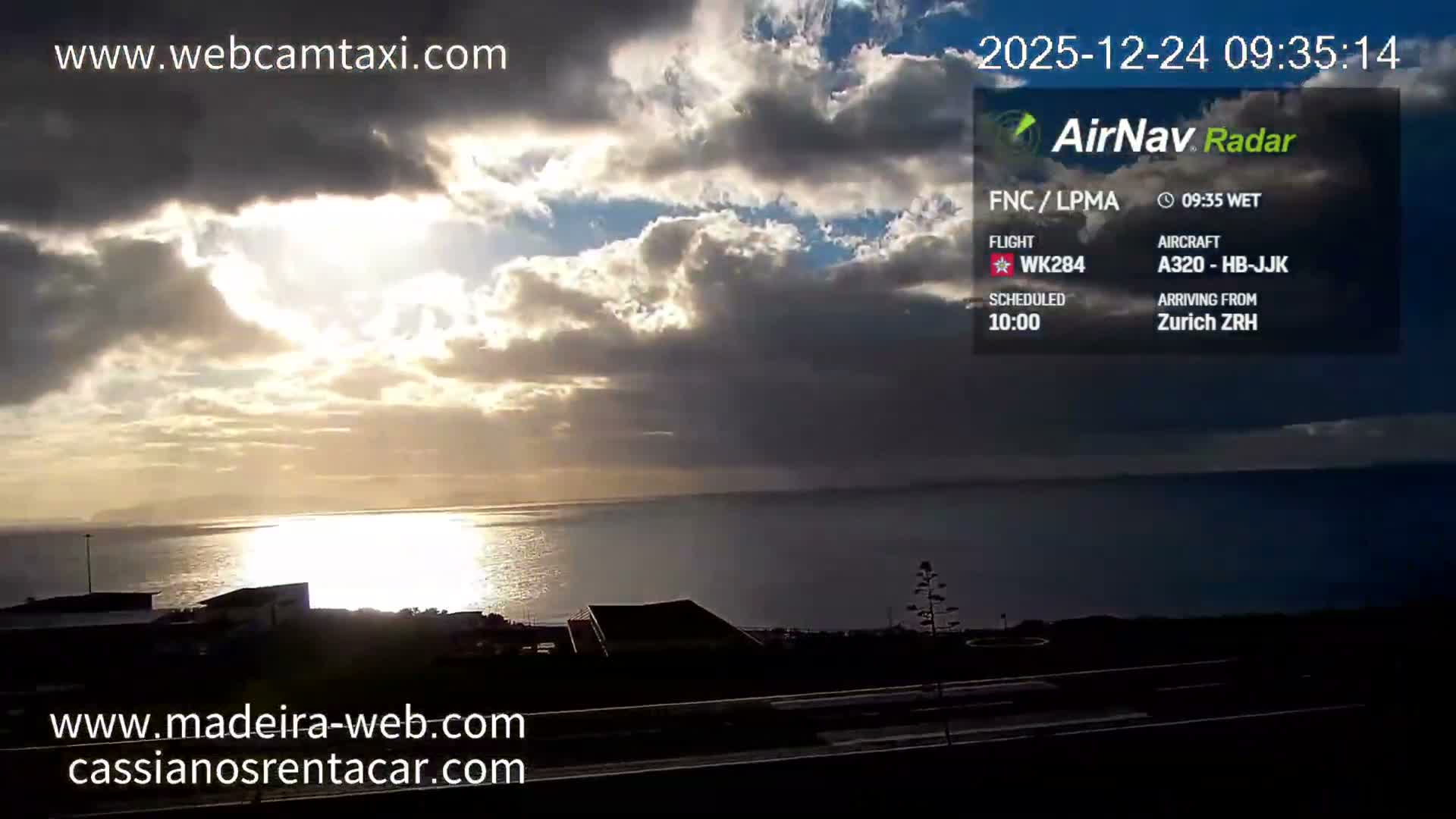 A bright and sunny morning with scattered clouds overlooks a shimmering sea reflecting the sun's glare, a distant coastline, and some foreground buildings.