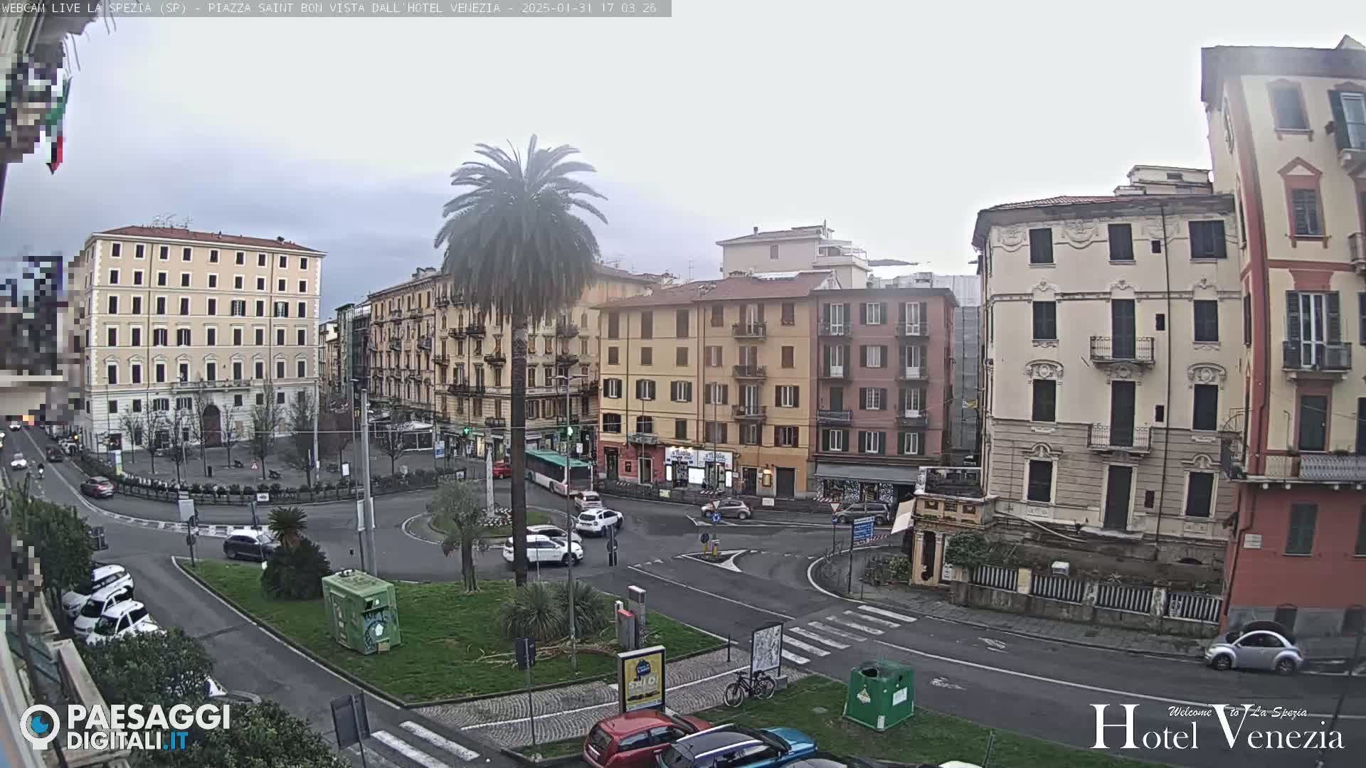 A city square with several multi-story buildings, a roundabout, cars, and a palm tree, under an overcast sky.