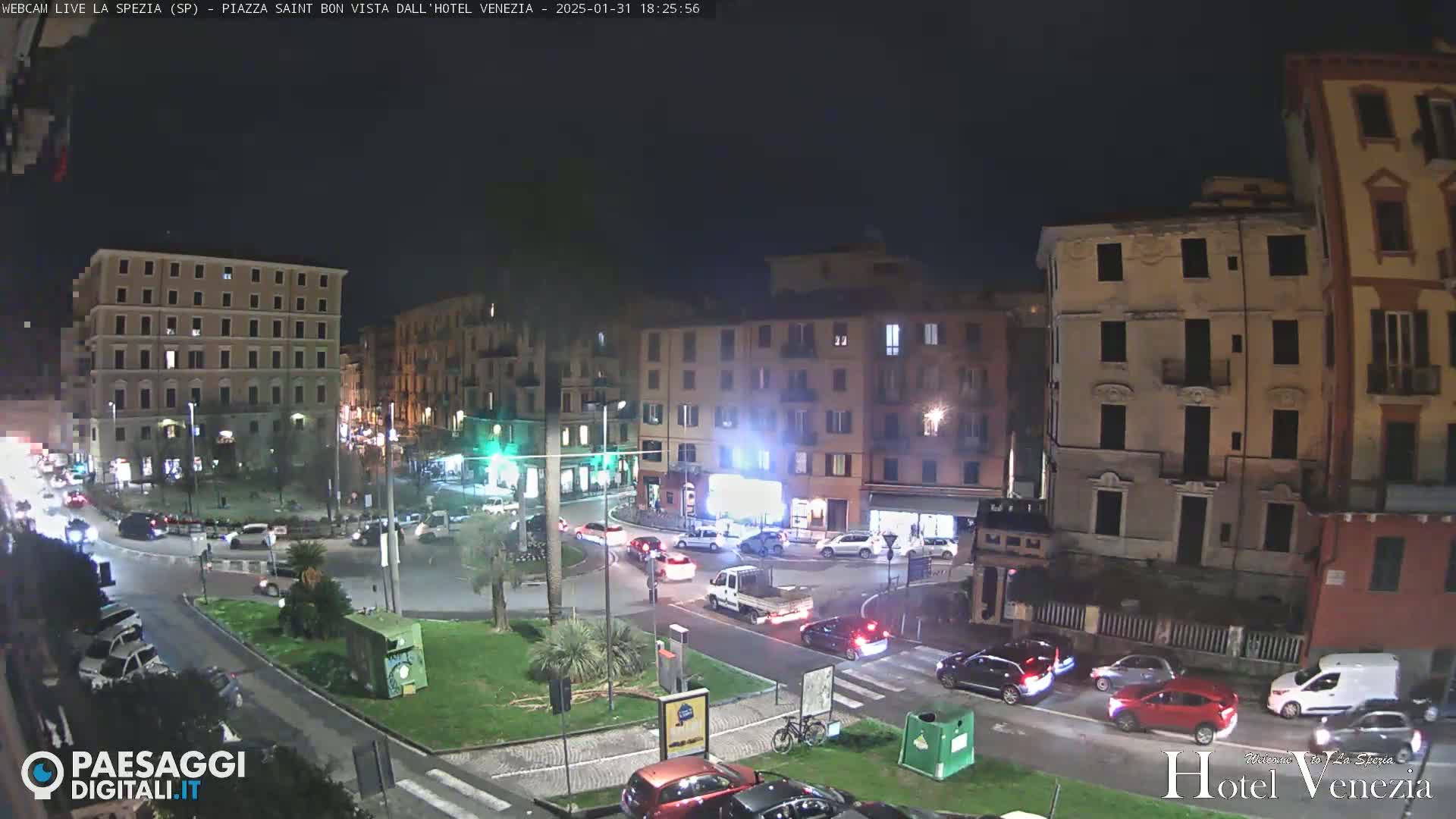 A nighttime, city-center roundabout scene shows several multi-story buildings surrounding a small park, with cars moving around the roundabout and parked along the streets.