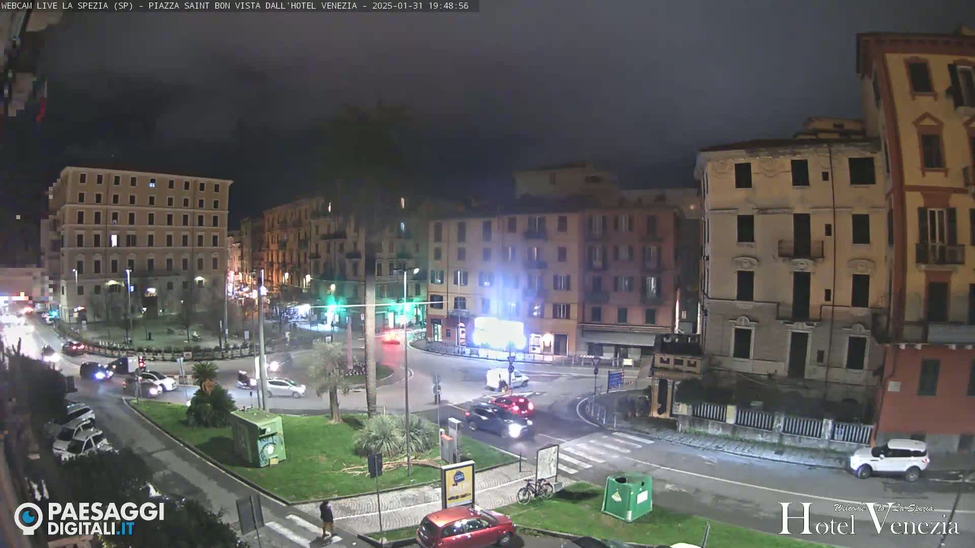 A nighttime view of a city square with cars and buildings under a dark, cloudy sky.