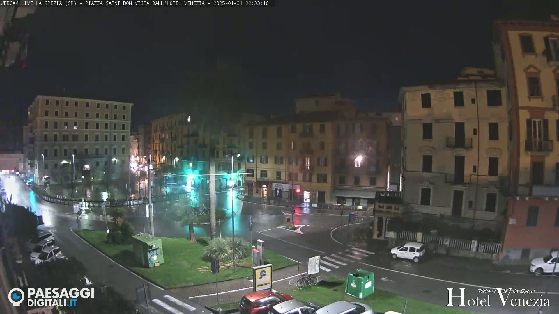 A rain-slicked nighttime city square, surrounded by multi-story buildings, with a few cars parked and driving.