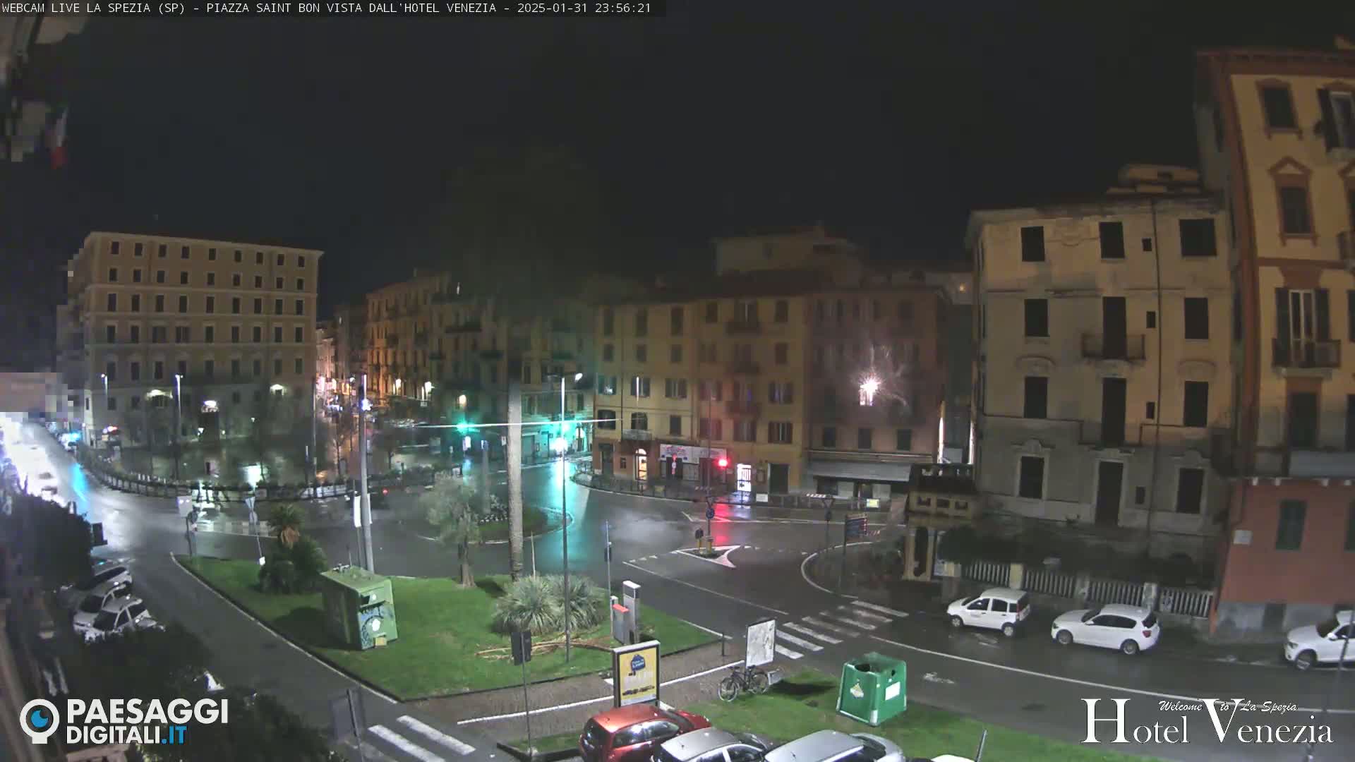 A nighttime view of a rain-slicked town square in a European city, surrounded by multi-story buildings with streetlights illuminating the wet streets and parked cars.