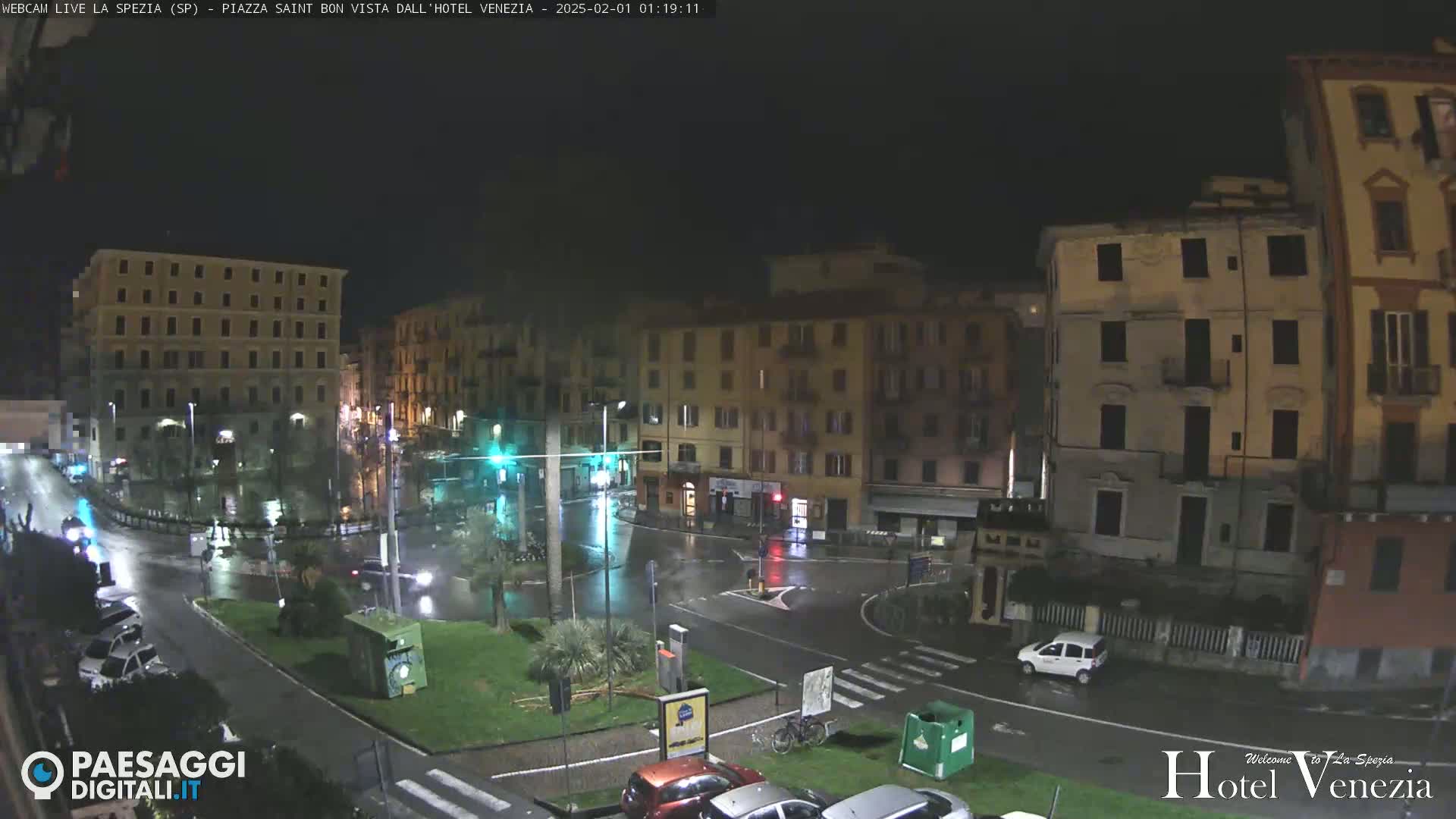 A rain-slicked town square at night, surrounded by multi-story buildings, with a few cars visible.