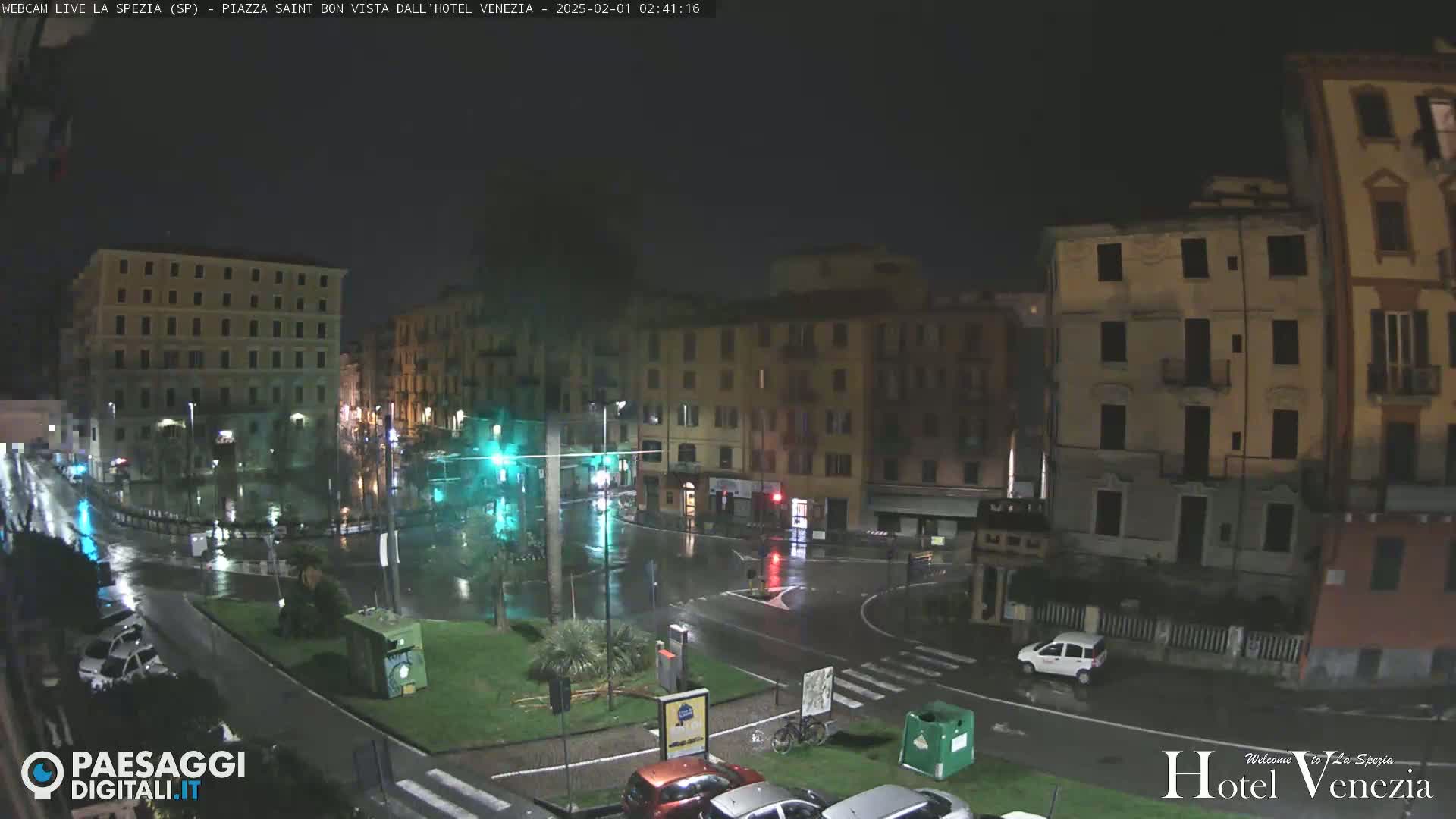 A nighttime view of a rain-soaked town square surrounded by multi-story buildings, with cars parked along the perimeter and a few driving through.