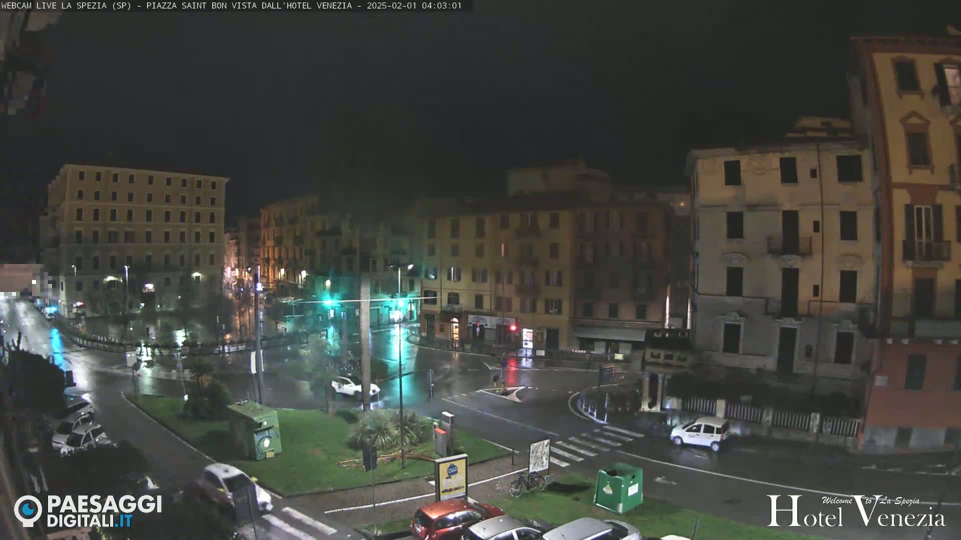 A rain-slicked city square at night, surrounded by multi-story buildings, with several cars visible on the wet streets.