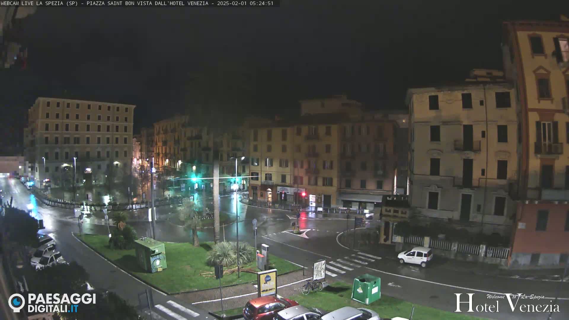 A rain-slicked town square at night, surrounded by multi-story buildings, with a few cars visible and some greenery in the center.