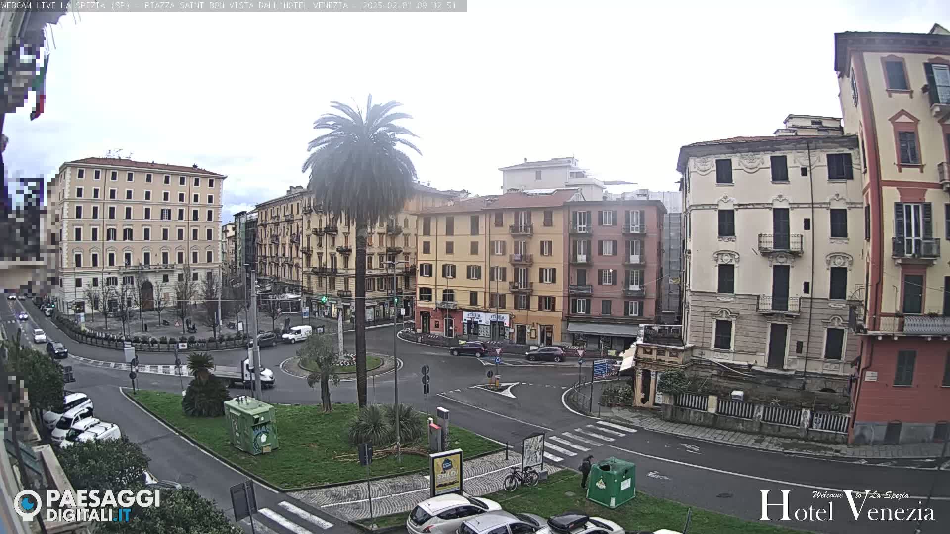 A mostly cloudy day reveals a city square with several multi-story buildings, a palm tree in the center, and cars parked along the perimeter.