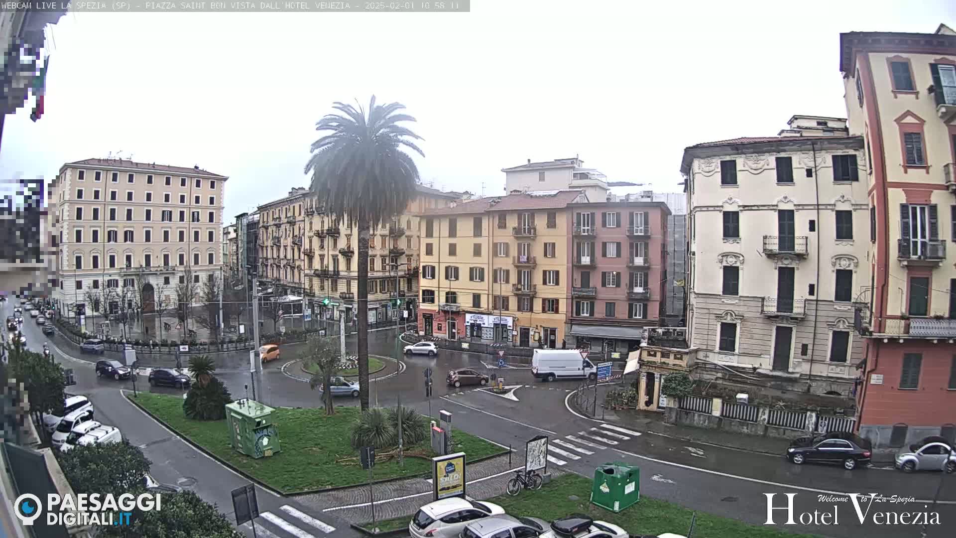 A wet, overcast city square is seen from an elevated viewpoint, showing several multi-story buildings, cars, a roundabout, a palm tree, and some parked bicycles.