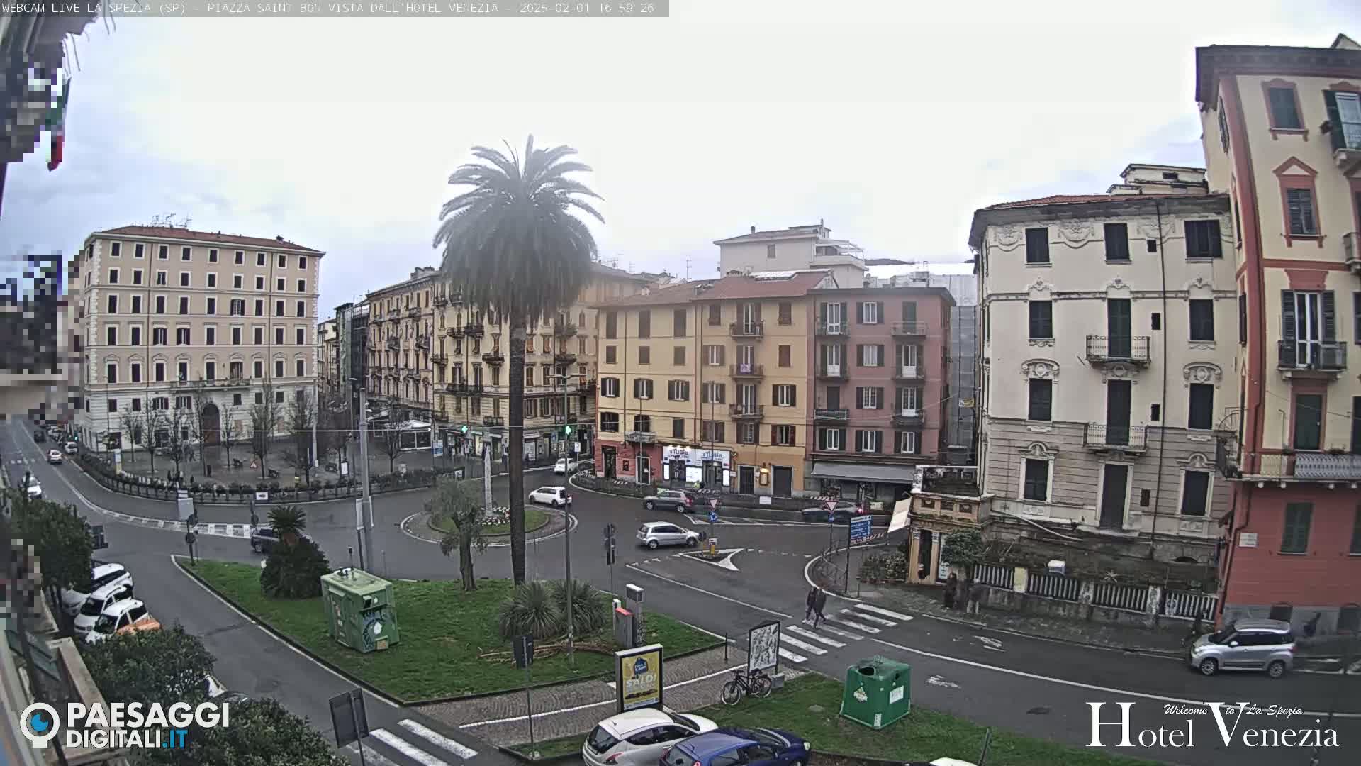 A cloudy day shows a city square with several multi-story buildings surrounding a circular traffic area with cars and a palm tree in the center.