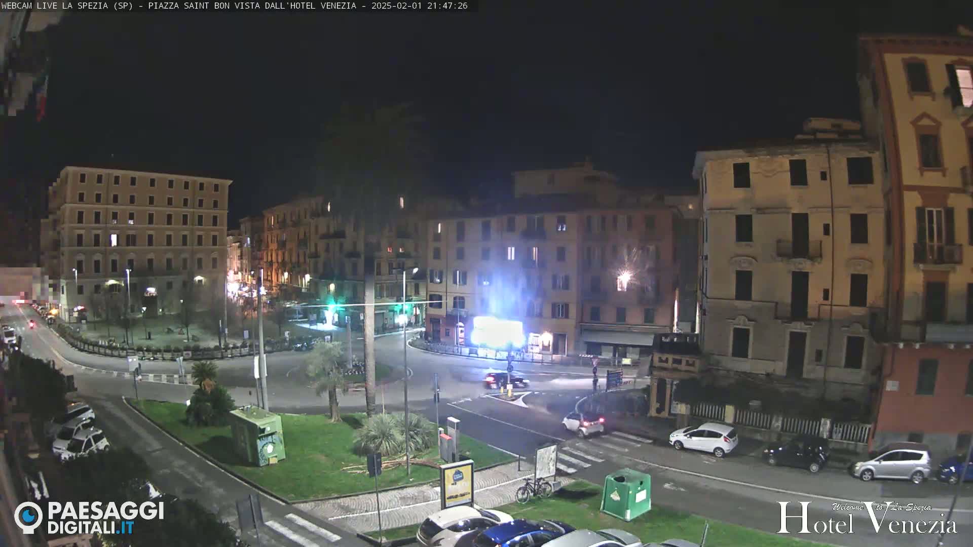 A nighttime view of a city square with several multi-story buildings, cars, and a small grassy area in the center.