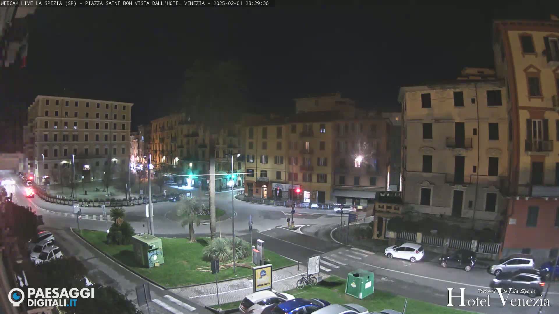 A nighttime view of a town square with several multi-story buildings surrounding a circular roadway, with cars parked and driving around the area.