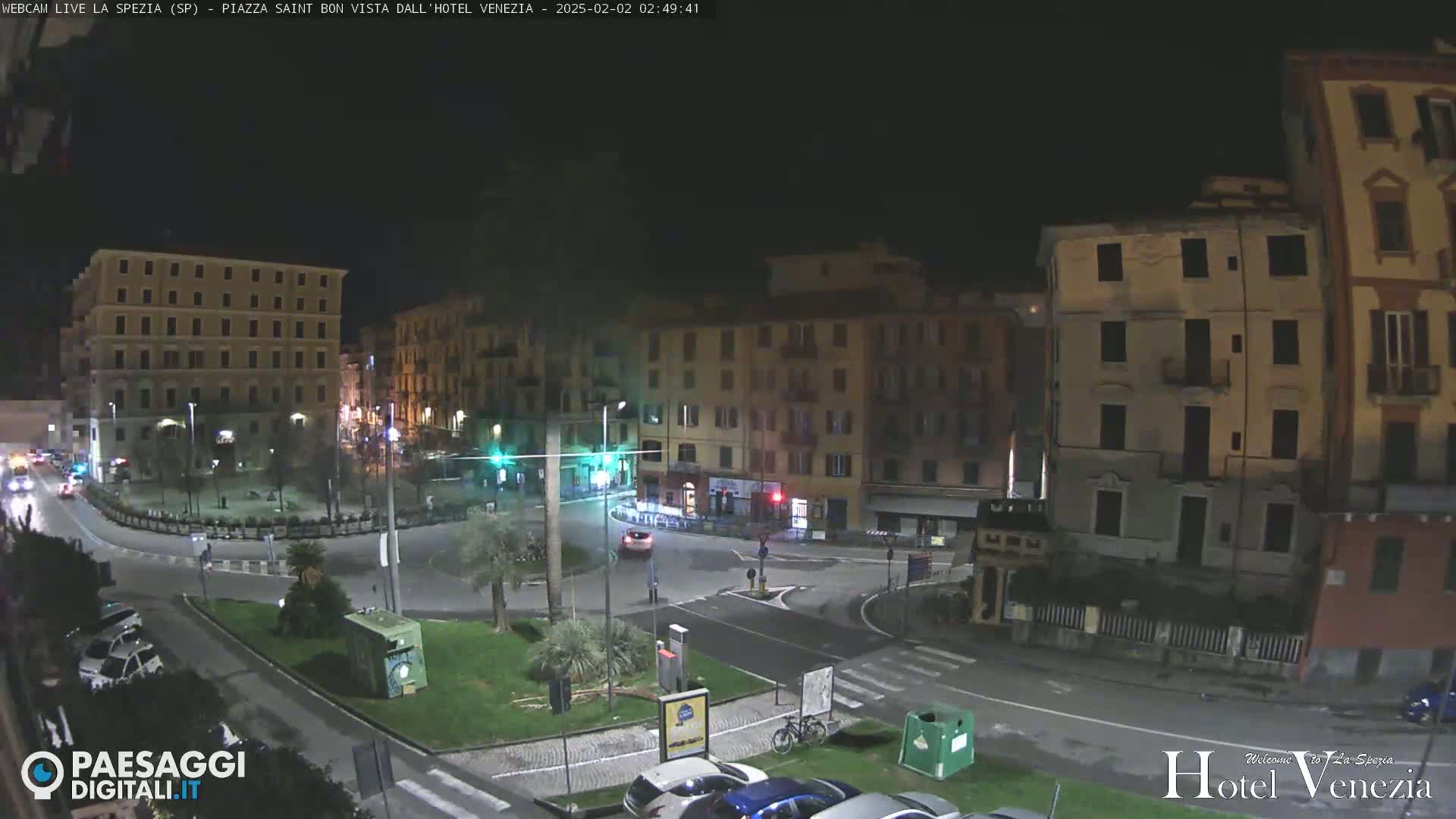 A nighttime view of a city square with several multi-story buildings, a roundabout, and parked cars.
