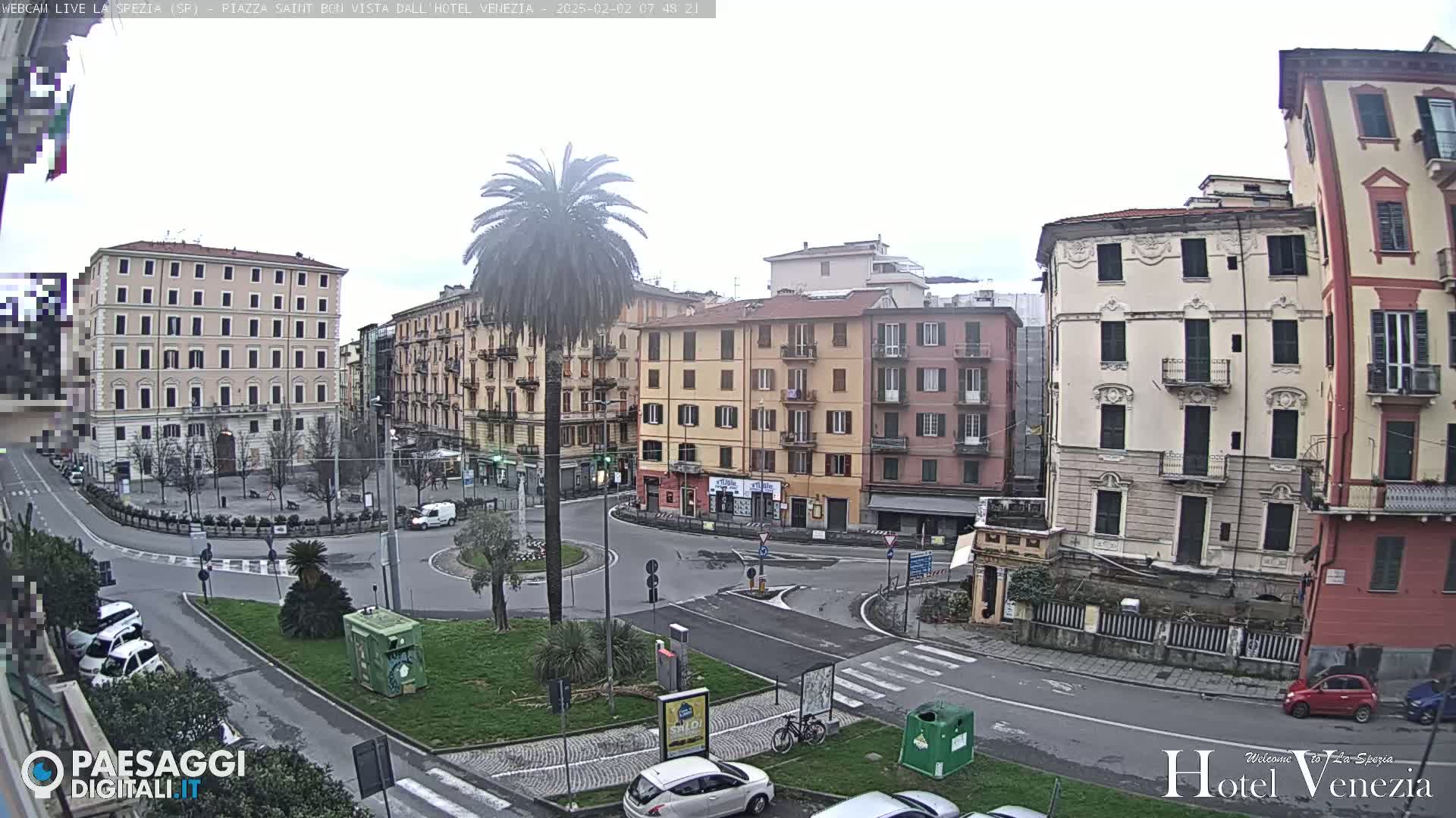 A mostly overcast day shows a traffic circle in a European city square surrounded by multi-story buildings, a palm tree, and several parked cars.
