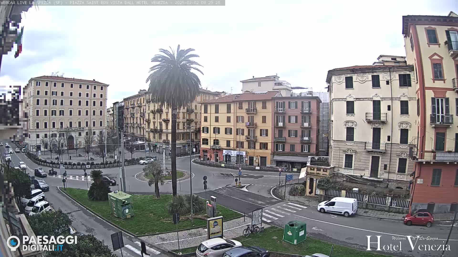 A mostly cloudy day reveals a European city square with multi-story buildings, a roundabout, and sparse traffic.