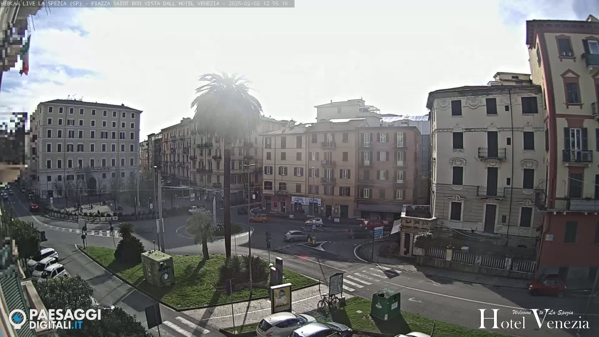 A hazy, sunny day overlooks a city square with several multi-story buildings, a roundabout, and a palm tree in the center.