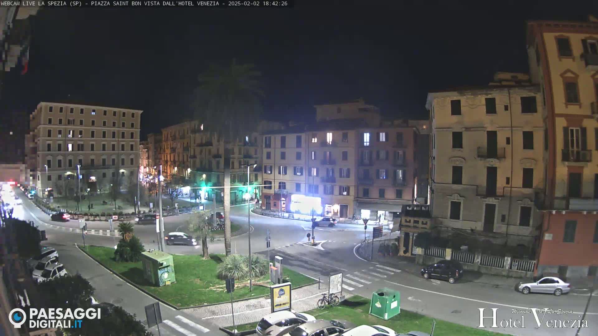 A nighttime view of a European city square with several buildings surrounding a roundabout, cars parked along the edges, and a few cars driving.
