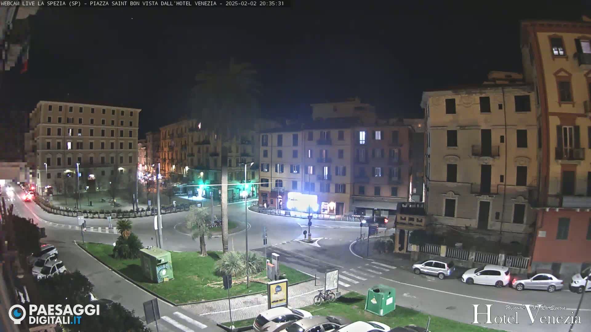 A nighttime view of a mostly empty plaza with several multi-story buildings surrounding it, a few cars parked and driving, and some greenery.