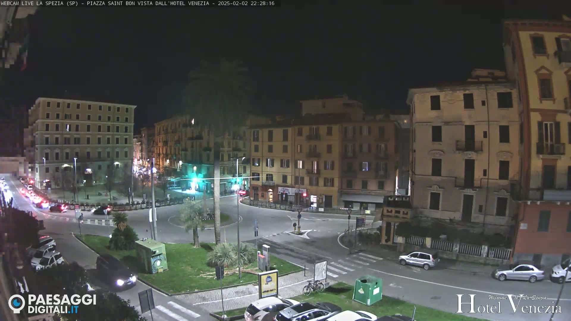 A nighttime view of a city square with several buildings, cars, and some greenery, under clear skies.