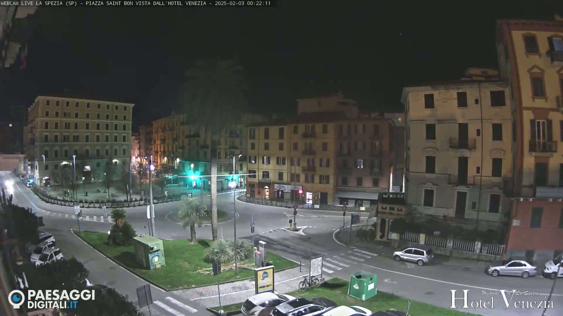 A nighttime view of a town square with several multi-story buildings surrounding a roundabout, some cars parked and driving, and a few trees and shrubs in the center.