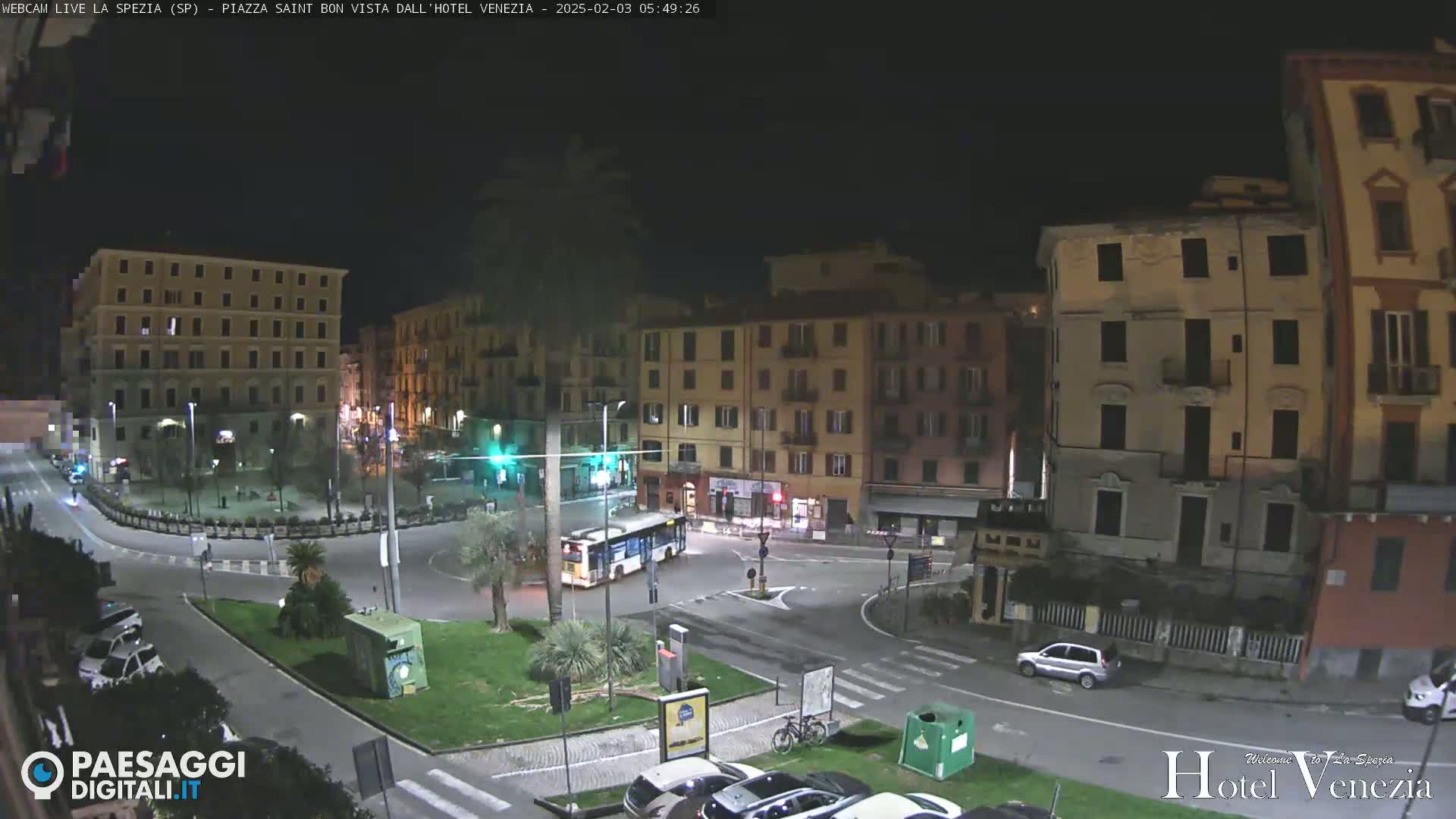 A nighttime, high-angle view of a city square with multi-story buildings surrounding a roundabout where a bus and several cars are parked.