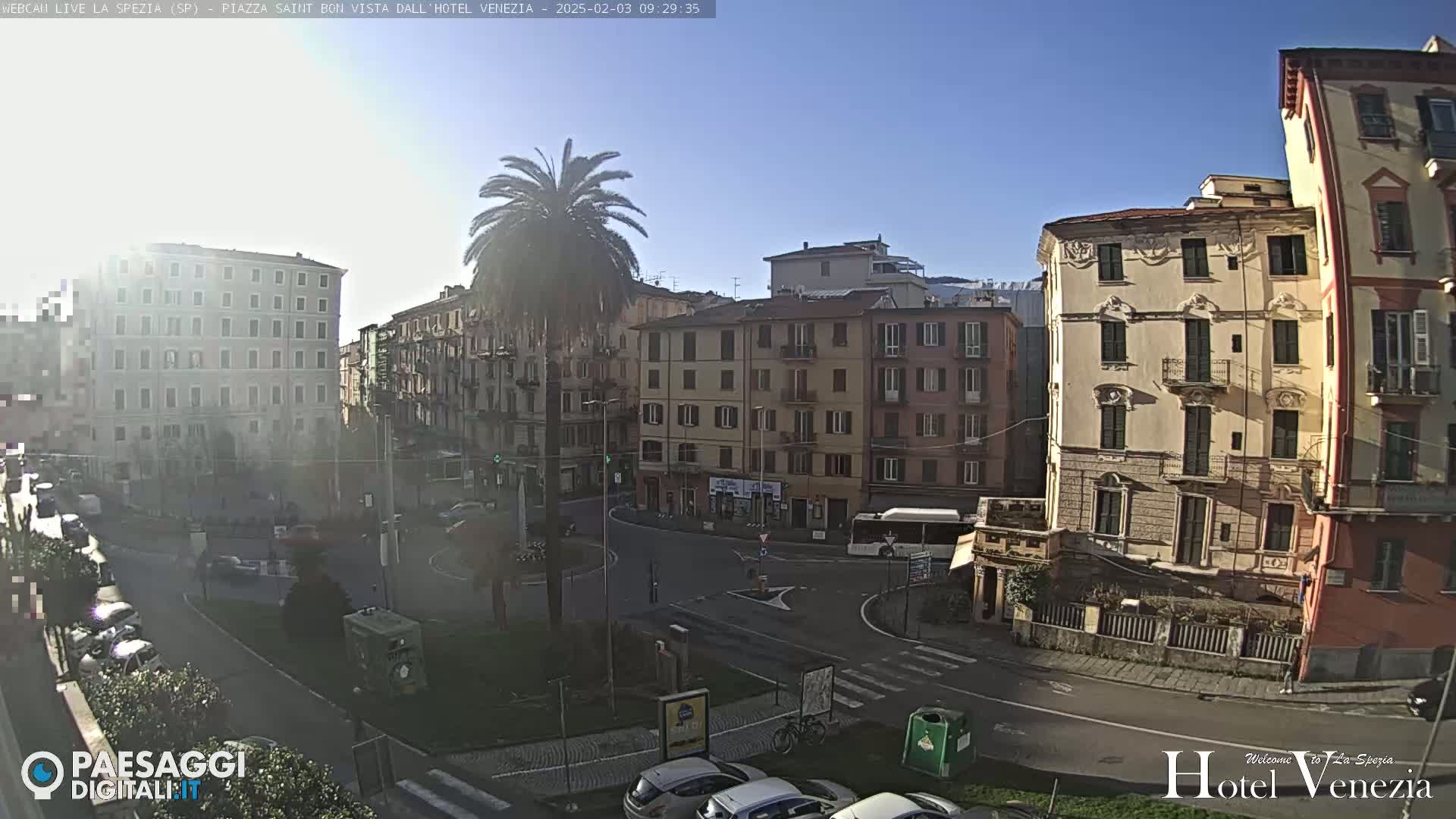 A sunny day in a European city square, showing several multi-story buildings and cars parked along a curved roadway.