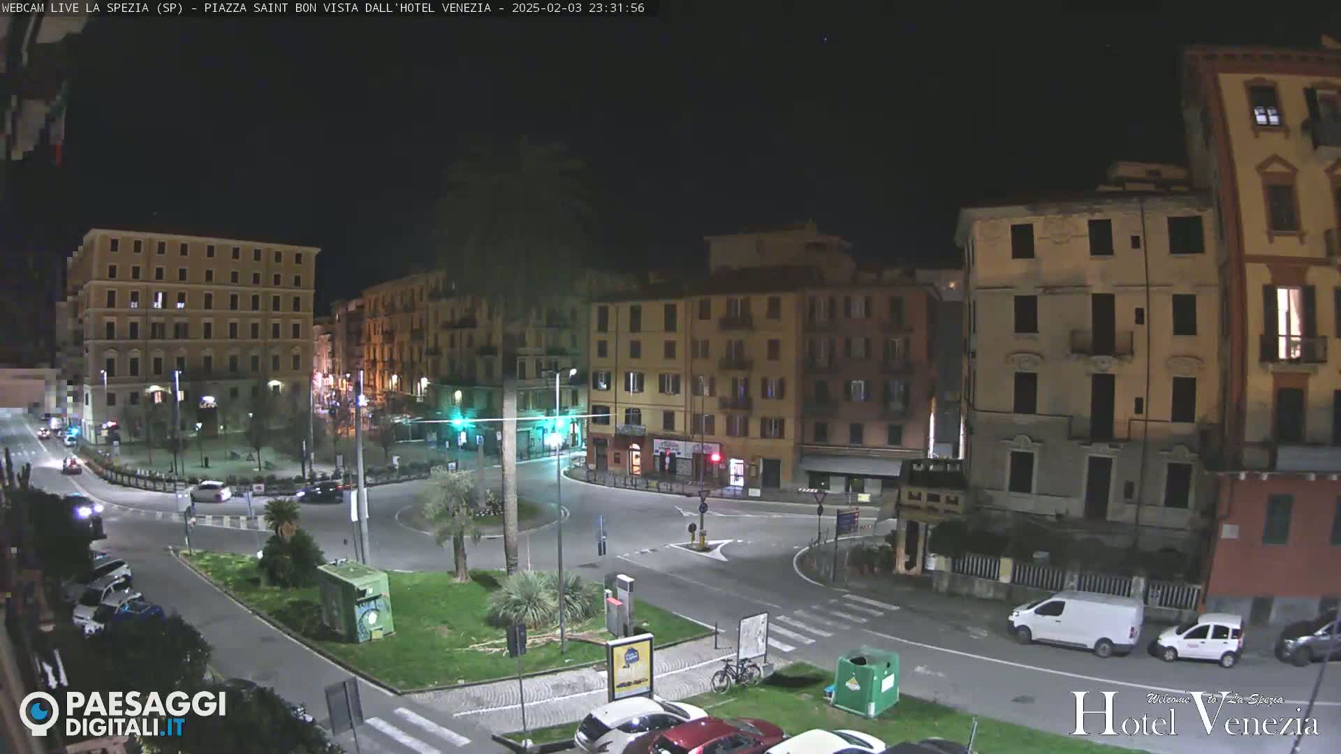 A nighttime view of a traffic circle in a city square, surrounded by multi-story buildings, with several cars parked and driving.