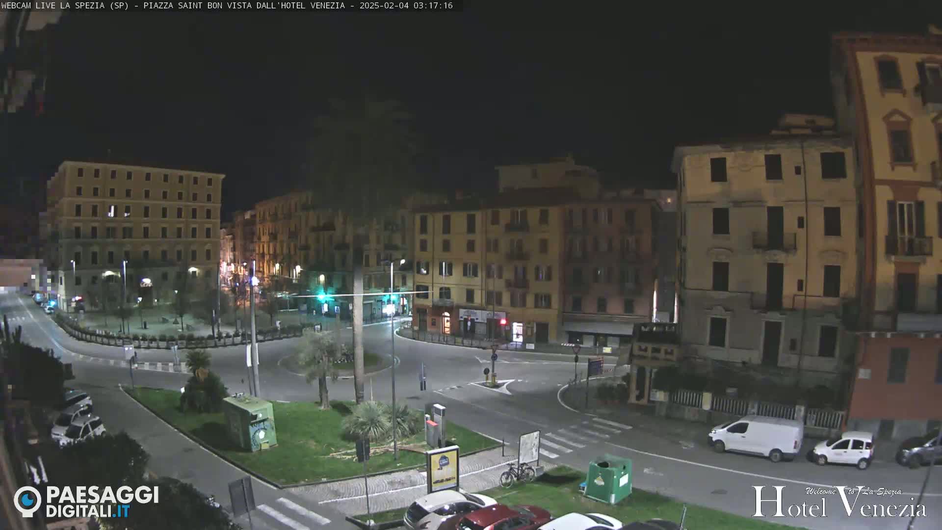A nighttime aerial view of a mostly empty town square with several multi-story buildings surrounding it, and a few vehicles parked or driving in the square.