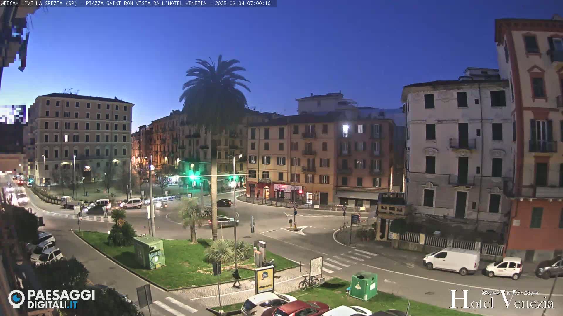 A twilight view of a city square with several multi-story buildings, a roundabout with cars, and palm trees, under a clear, dark-blue sky.