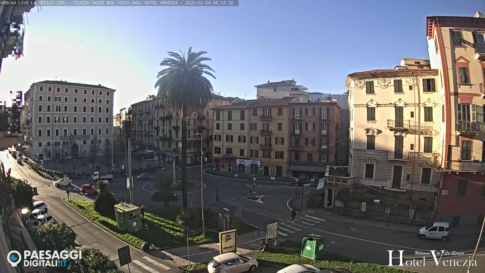 A sunny day reveals a city square with several multi-story buildings, a central palm tree, and a few cars in the surrounding streets.