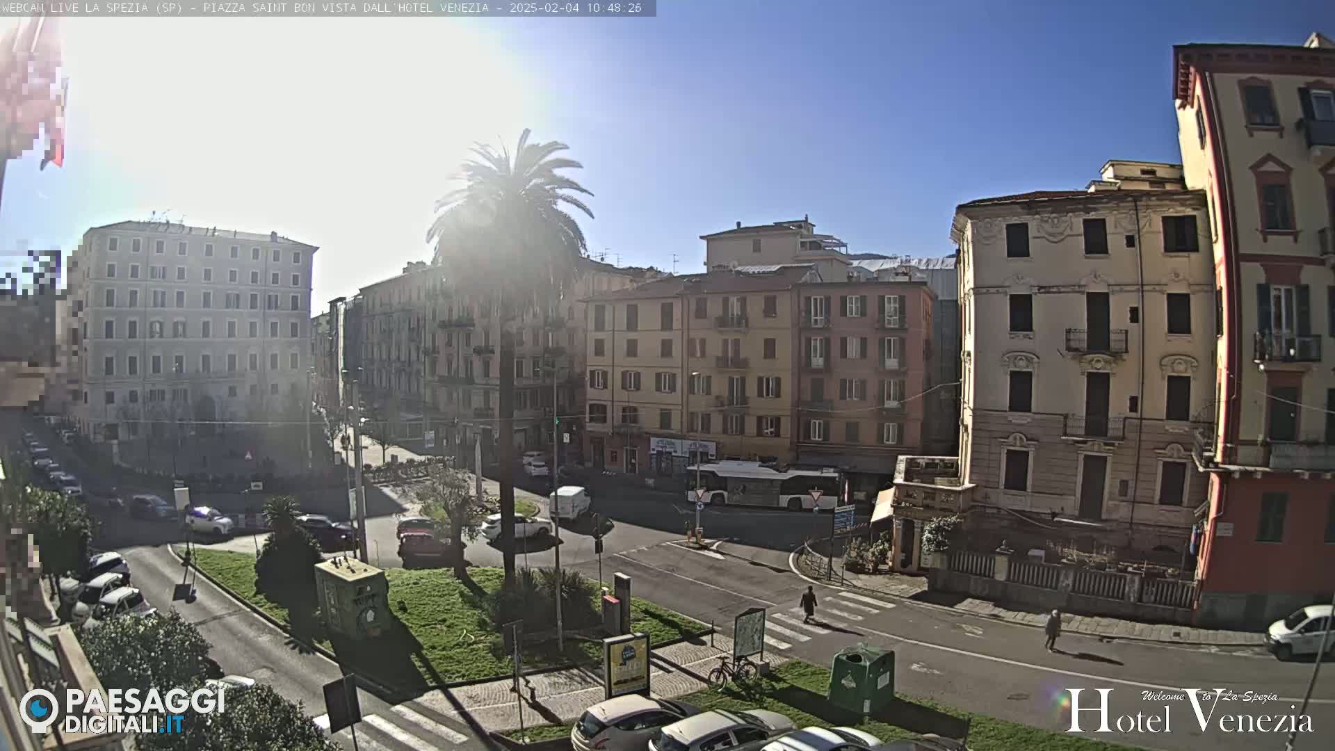 A sunny day reveals a European city square with several multi-story buildings, cars parked along the streets, and a few pedestrians walking.