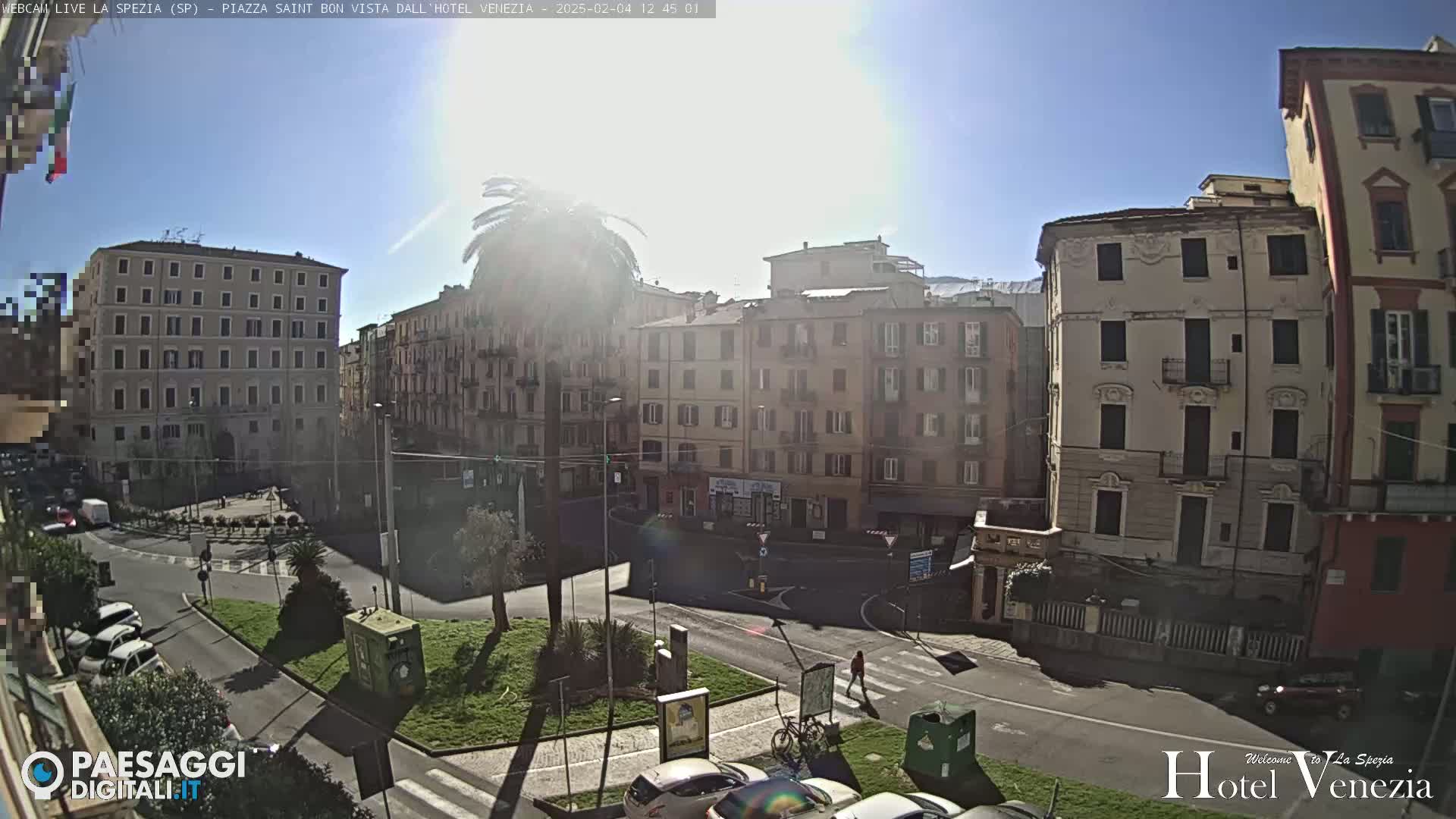 A sunny day reveals a city square with several multi-story buildings surrounding it, cars parked along the road, and a lone person walking across a crosswalk.