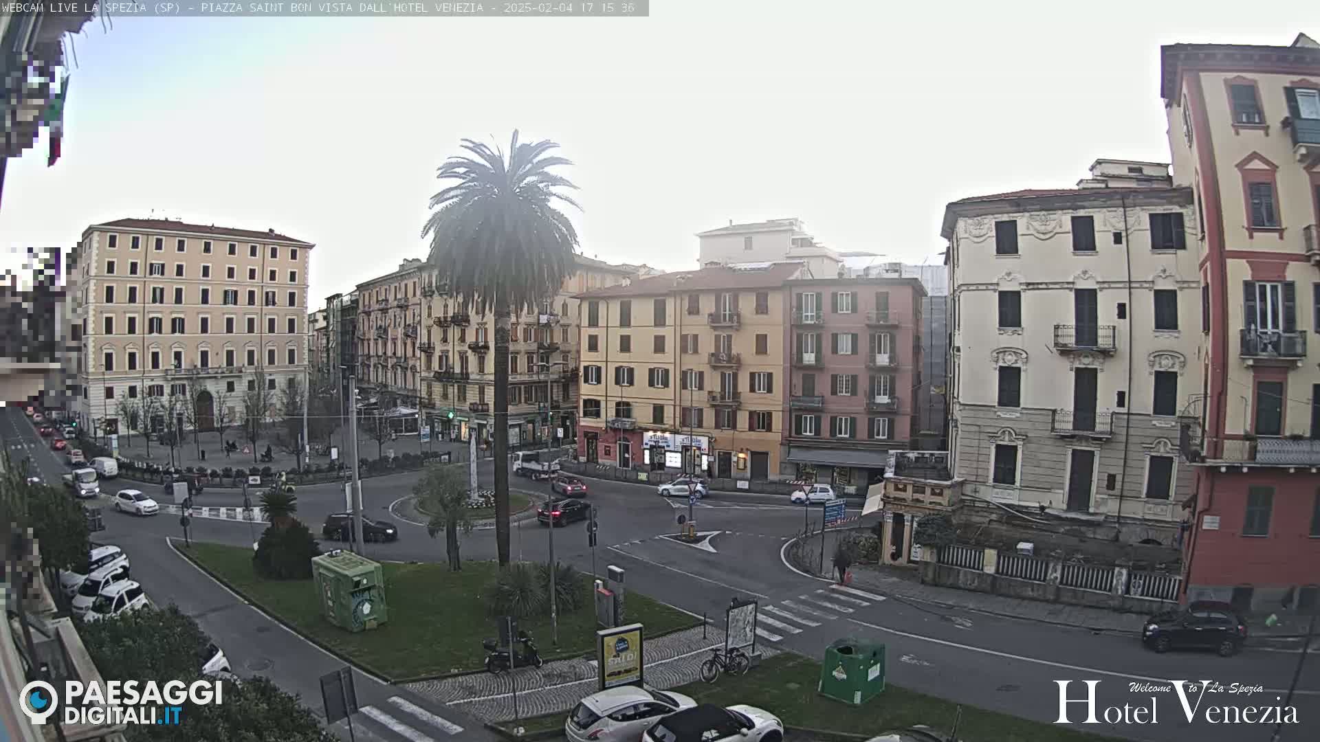 A mostly cloudy day reveals a city square in Italy, surrounded by multi-story buildings, with cars and a few pedestrians visible in a roundabout.
