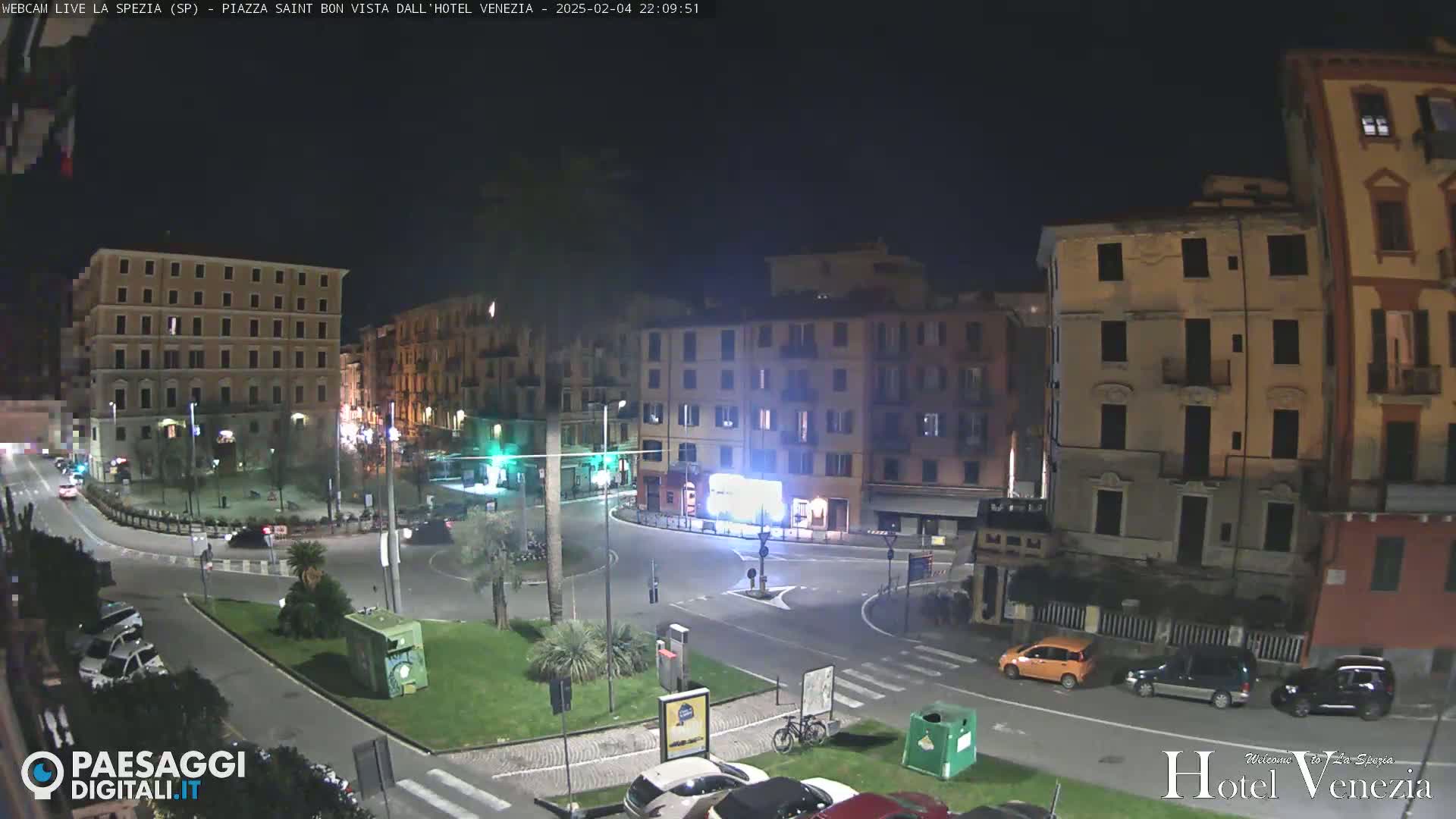 A nighttime view of a city square with several multi-story buildings surrounding it, some cars parked and driving, and a few palm trees in a small grassy area.