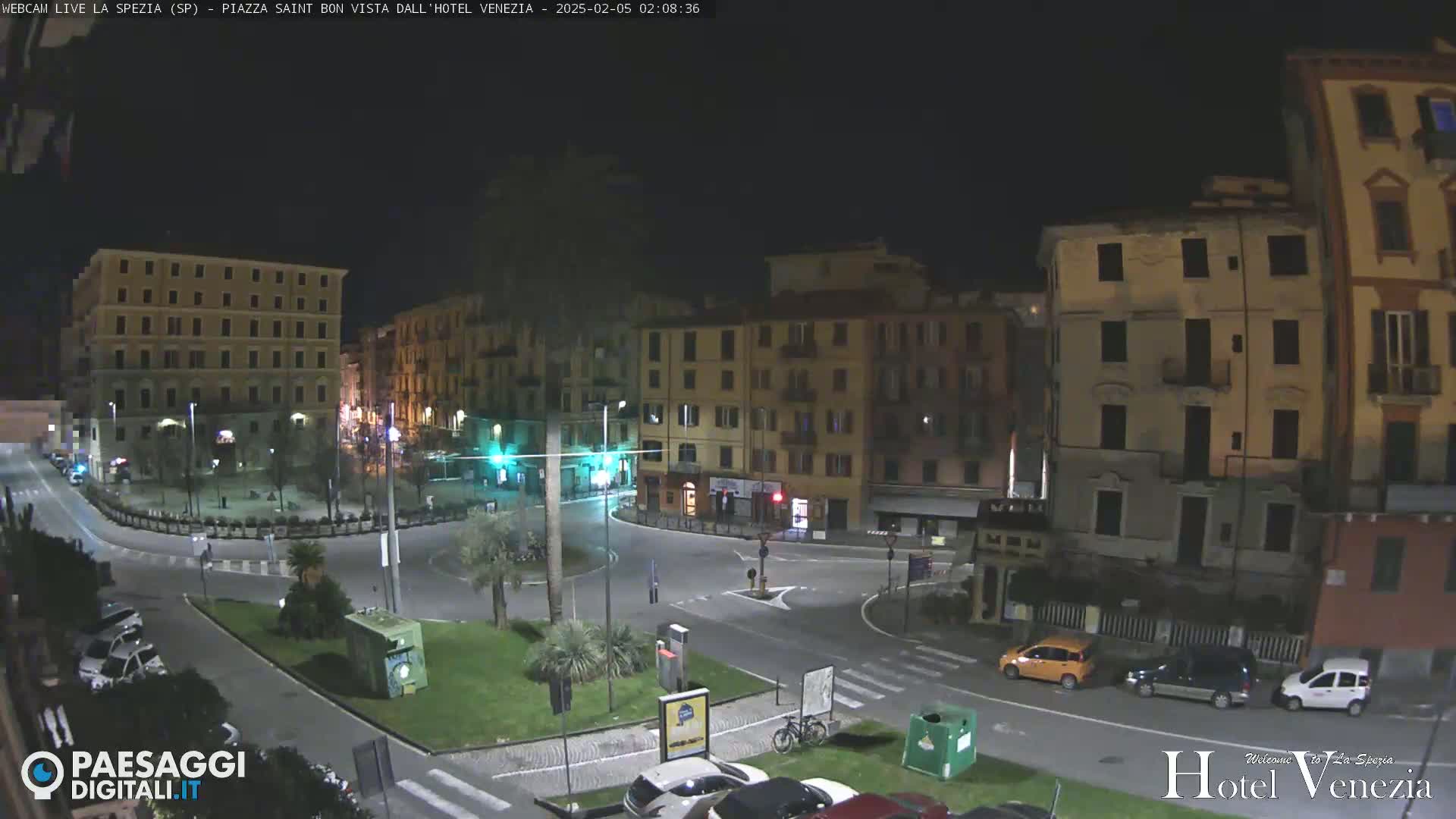 A nighttime view of a town square with several multi-story buildings surrounding a traffic circle, with cars parked along the perimeter and a few cars driving through.