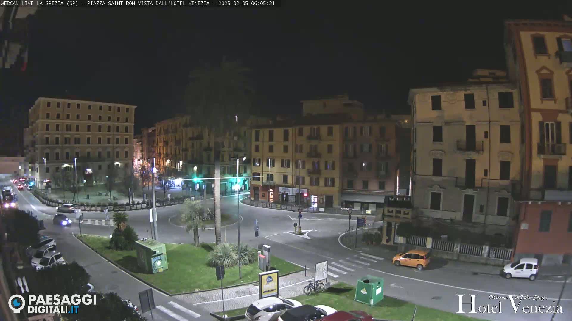 A nighttime view of a city square with several multi-story buildings surrounding a circular road, with a few cars and a bicycle present.
