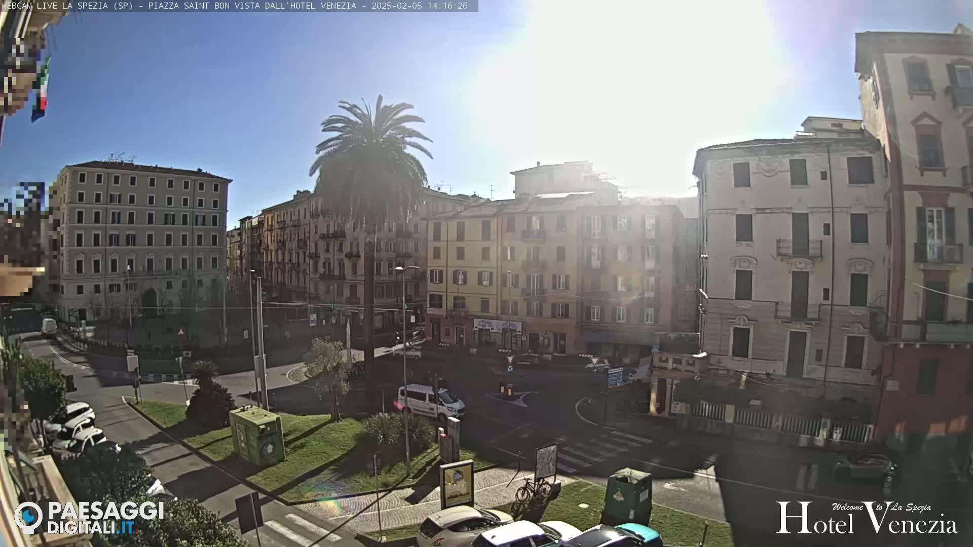 A sunny day reveals a city square surrounded by multi-story buildings, with cars parked and a few people visible.
